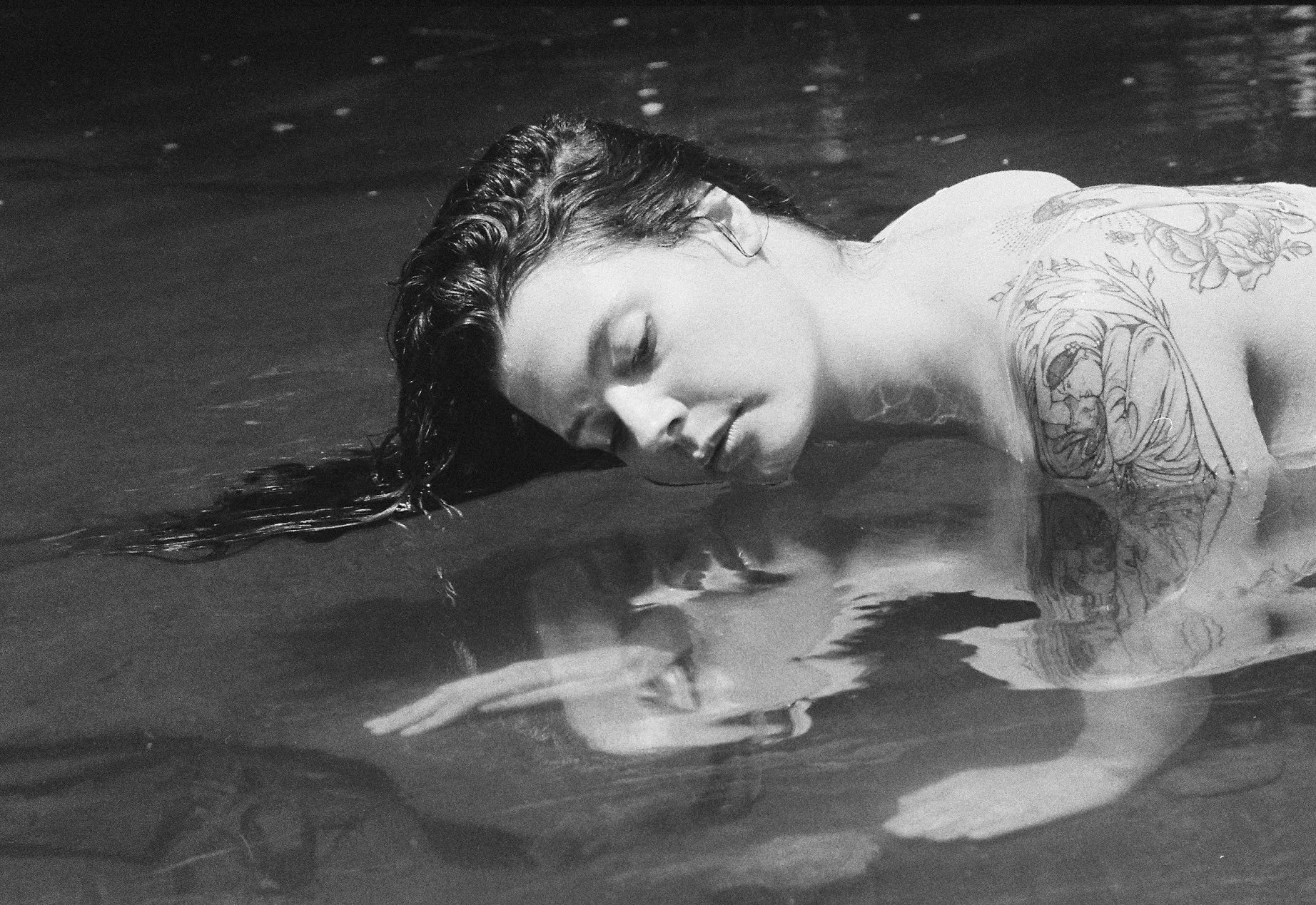 A black and white photo of a woman with long, wet hair floating on water, her eyes closed, and her face resting on the surface. She has tattoos on her shoulder and arm.
