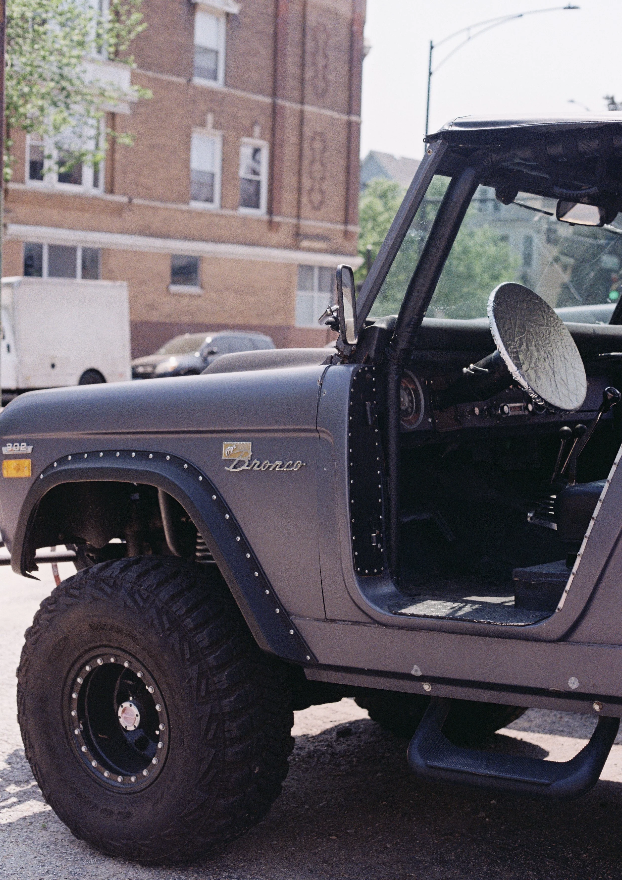 Close-up of a gray vintage Ford Bronco SUV with large off-road tires, parked on a city street with brick apartment buildings in the background.