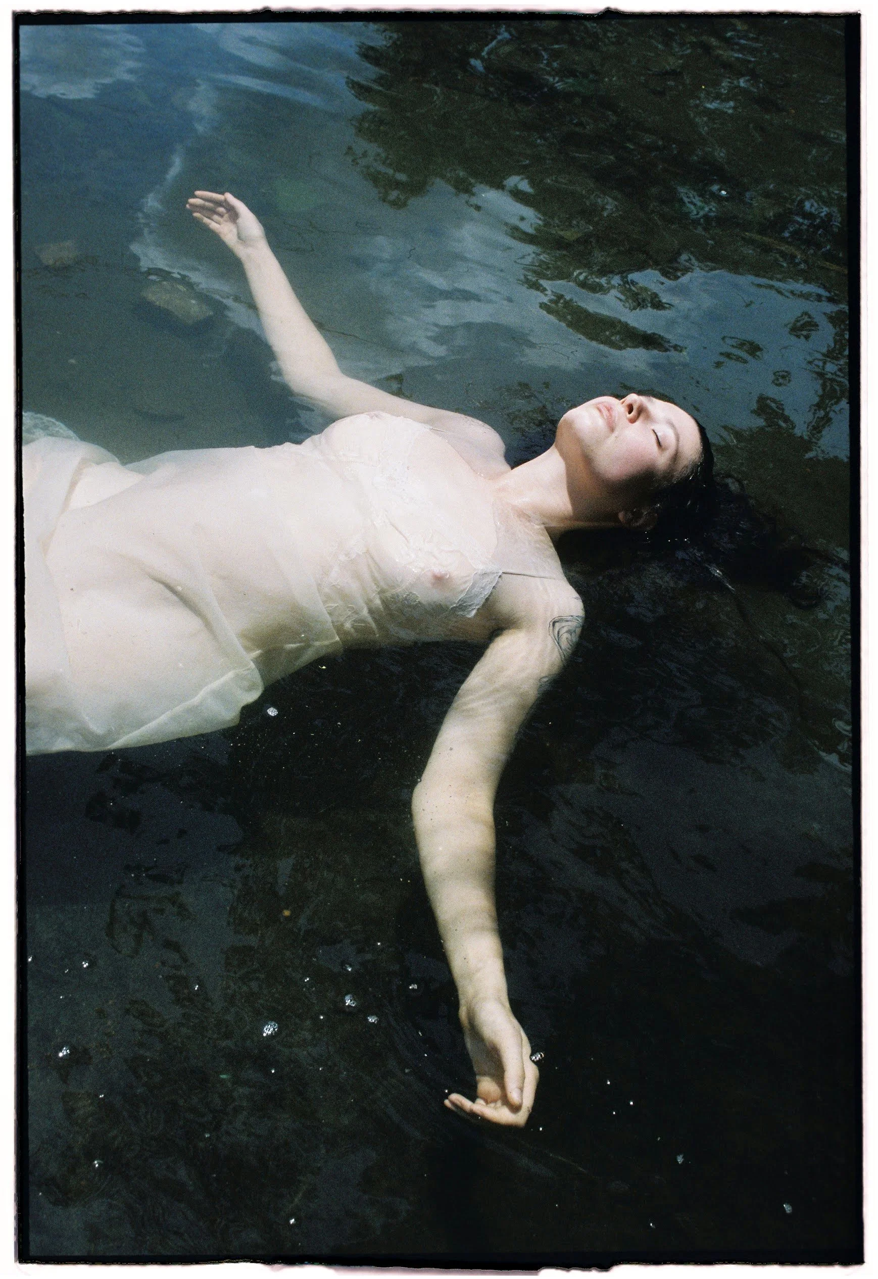 A woman floating in water with her eyes closed, wearing a light-colored dress, dark hair spread out and partially submerged.