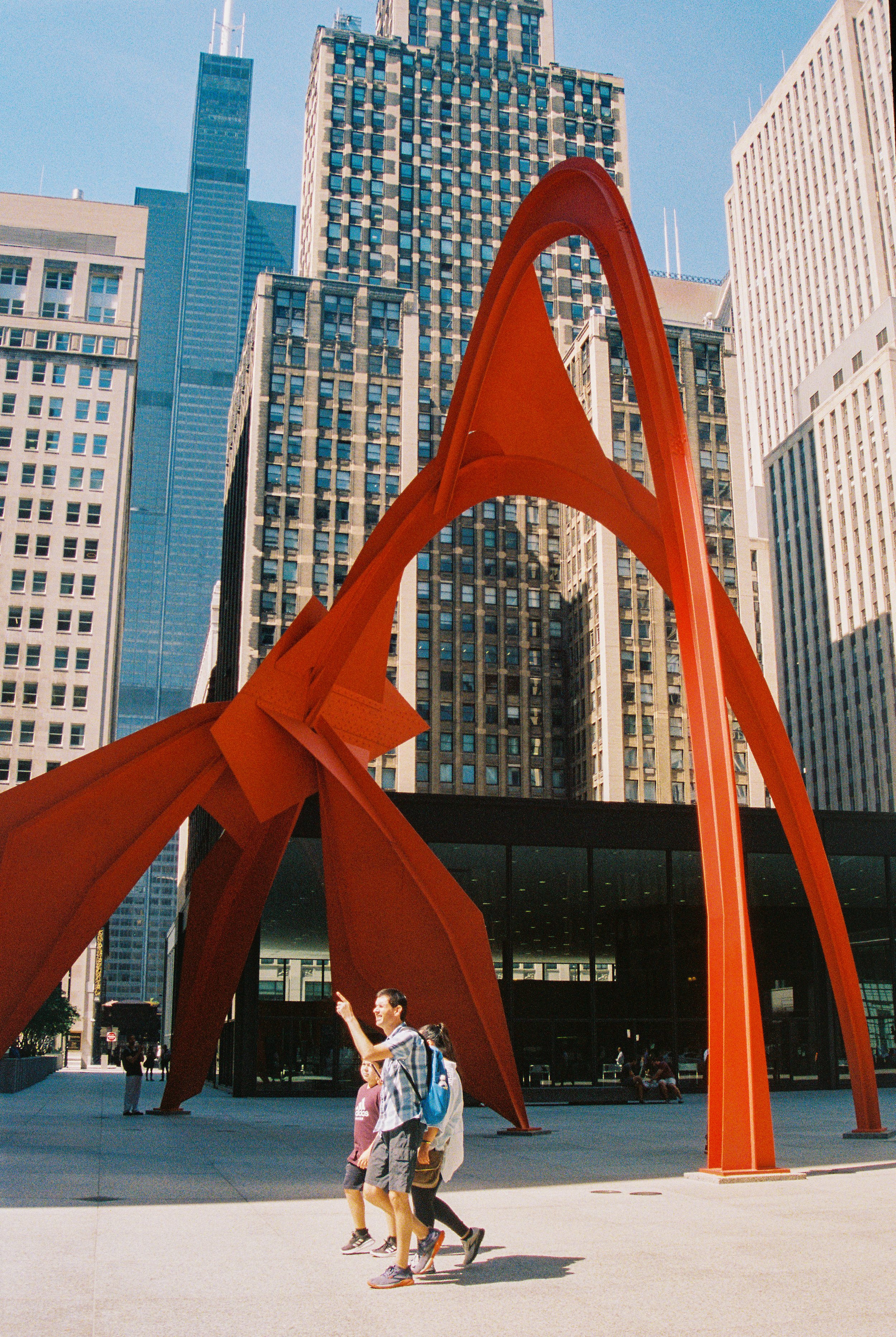 People walking in front of the Flamingo sculpture by Alexander Calder in an urban area surrounded by tall skyscrapers.