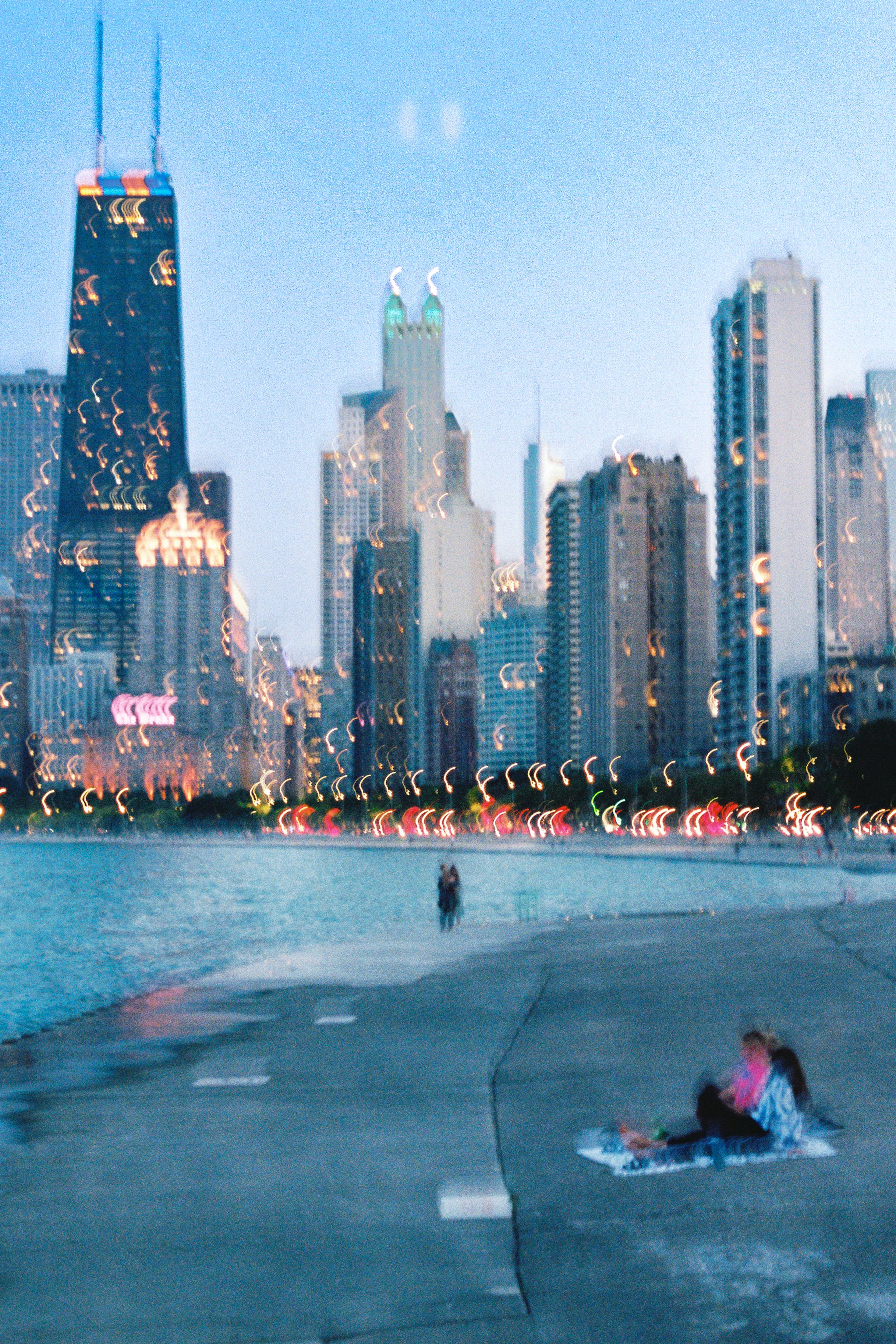 City skyline with tall buildings and a waterfront, with two people sitting on a blanket near the water's edge, and a person walking by in the background during twilight.