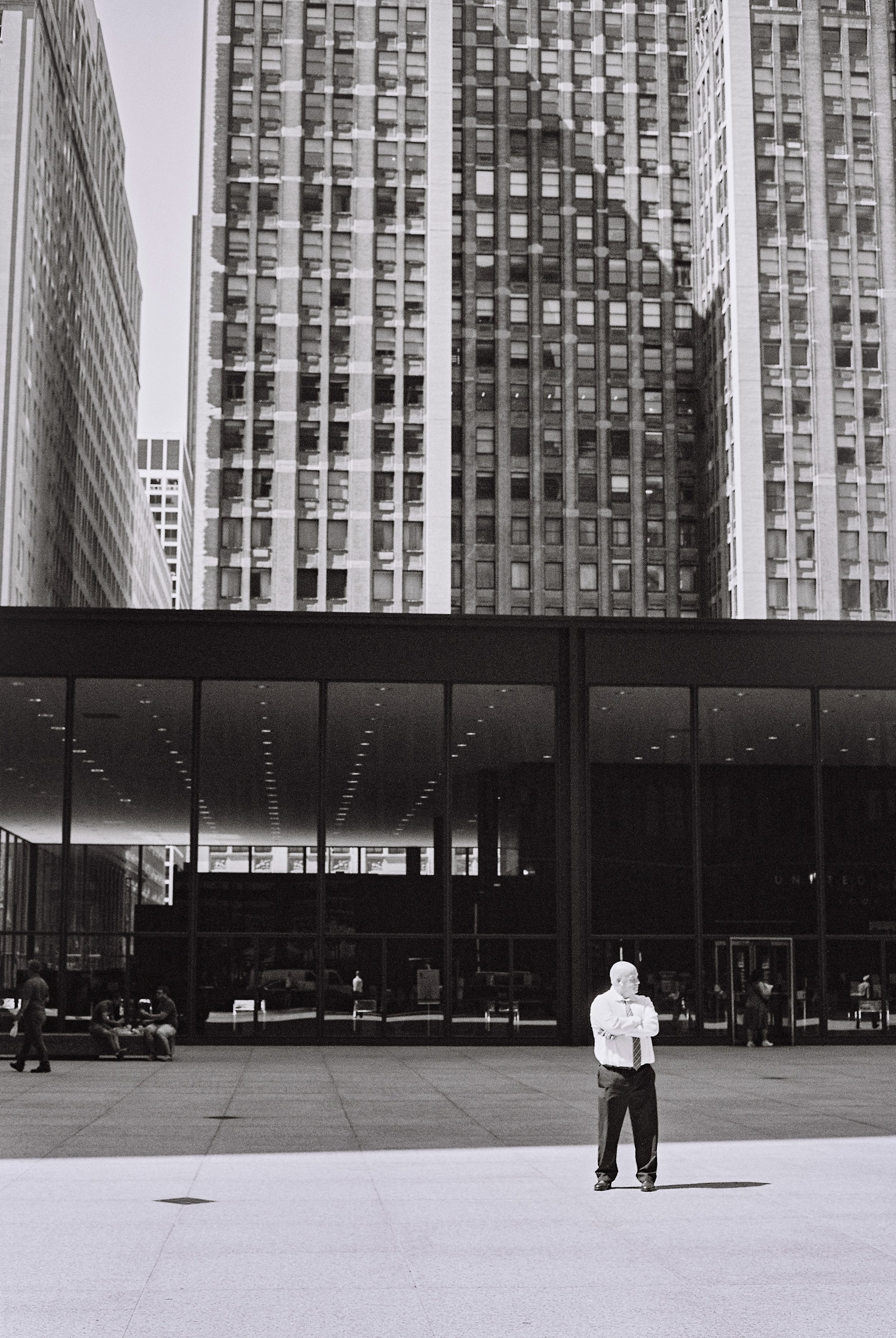 A man in a white shirt and tie standing with arms crossed in an open urban plaza, with tall office buildings in the background and glass building facade reflecting the interior lighting.