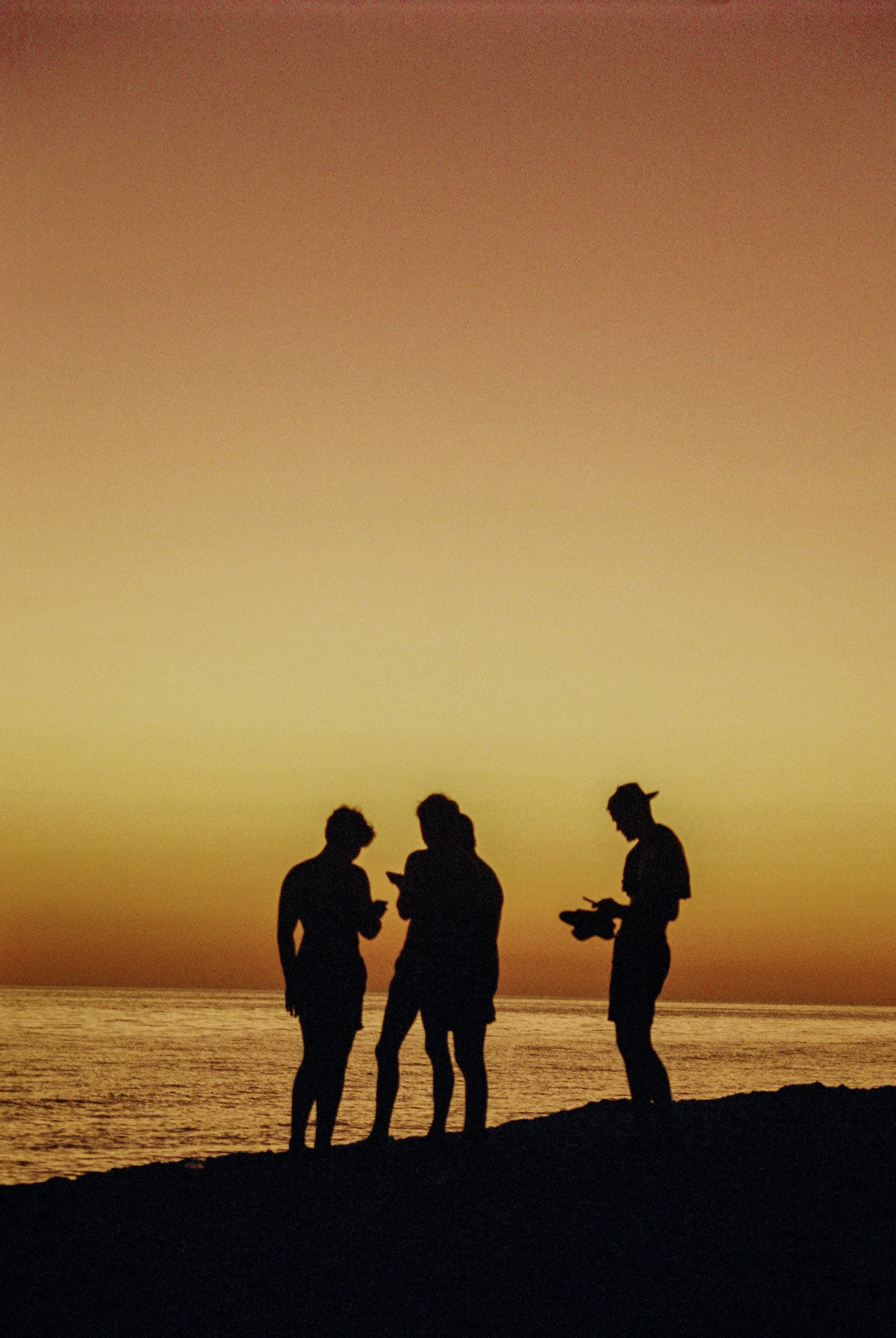 Silhouettes of four people standing on a beach at sunset, looking at their phones.