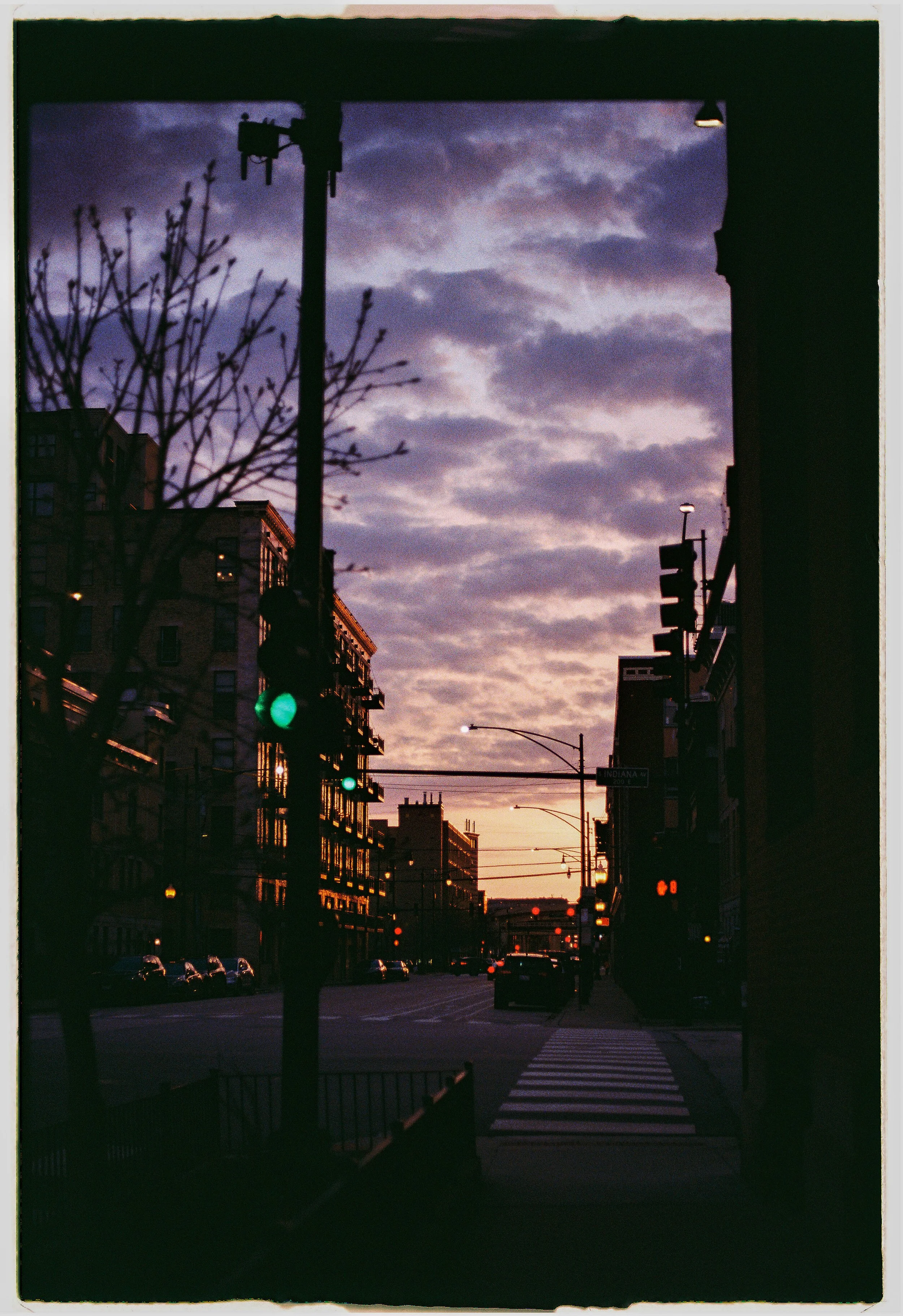 City street view during sunset with buildings, trees, cars, traffic lights, and a sky with clouds.