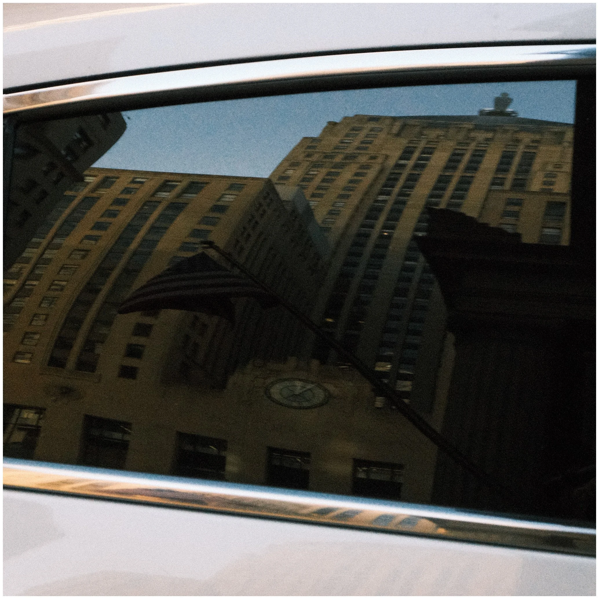 Reflection of tall city buildings on the dark glass window of a car, including a clock on one building and a flag partially visible with a striped pattern.