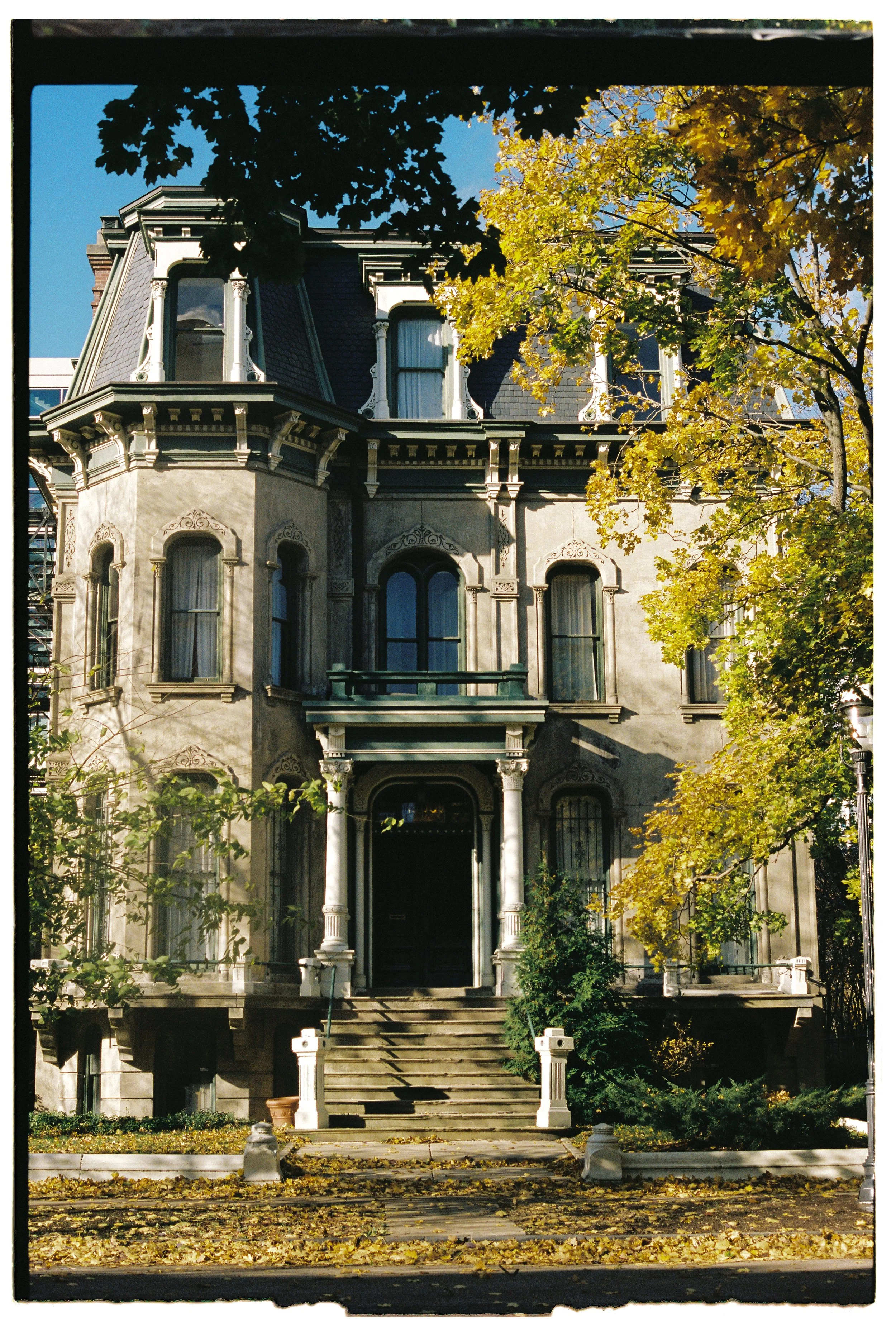 A tall, old Victorian-style house with intricate architectural details, multiple windows, and a front staircase leading to a black door, surrounded by trees with yellow leaves and fallen leaves on the ground, under a clear blue sky.