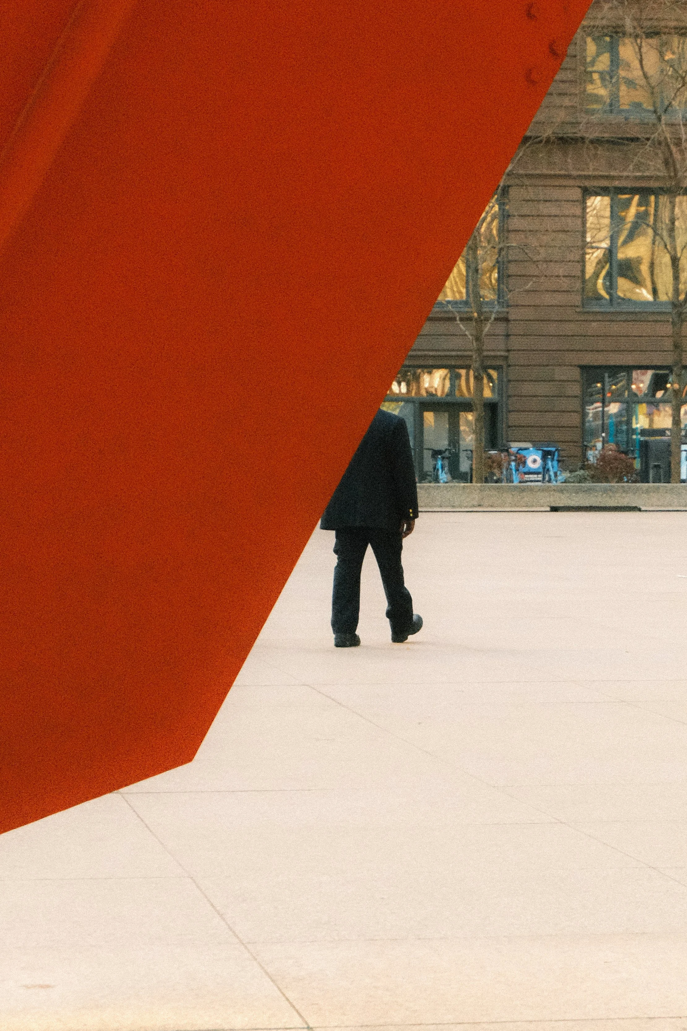 A person in dark clothing walking outside on a concrete area with modern buildings and trees in the background, partially obscured by a large red flamingo sculpture by Alexander Calder in Chicago.