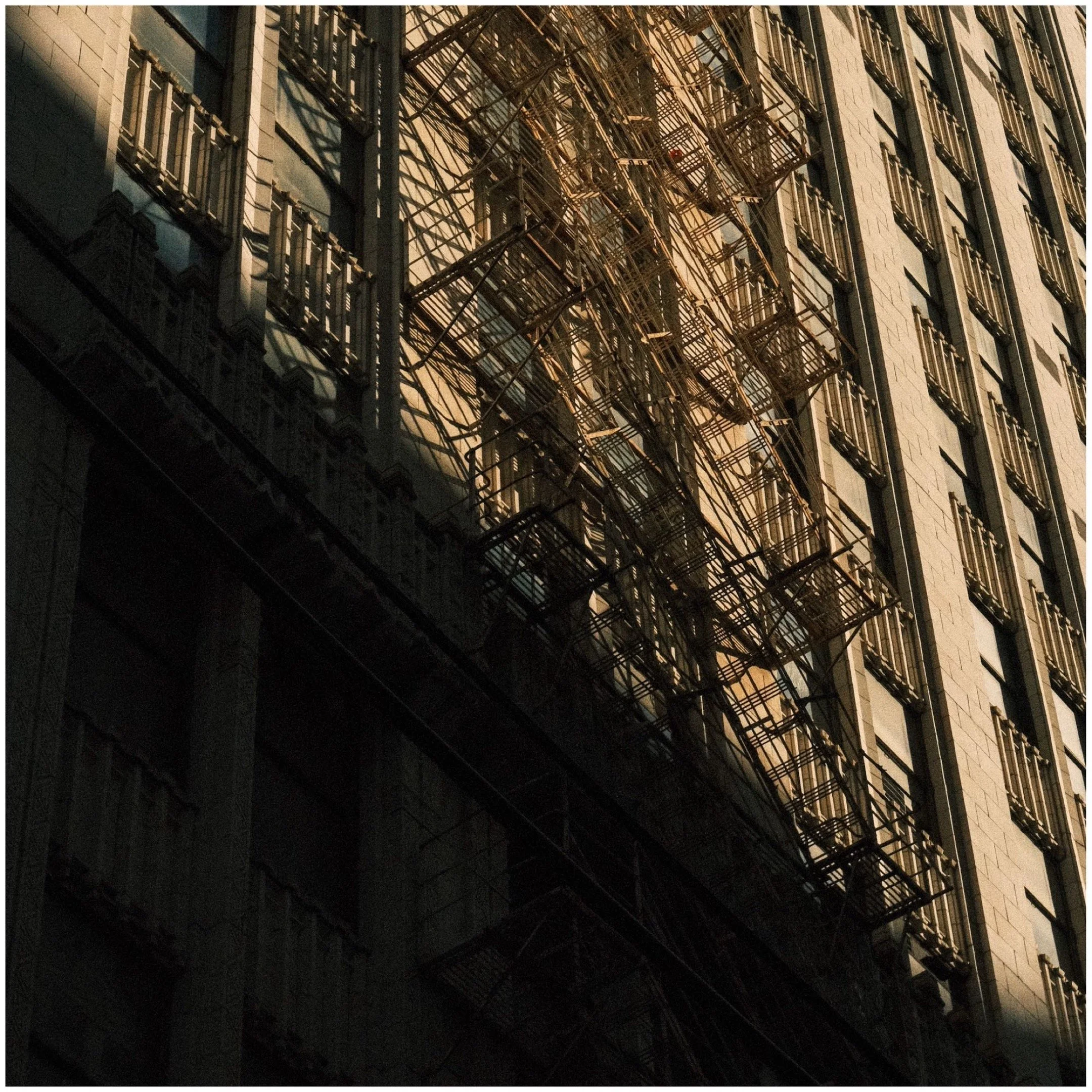 Close-up of a tall building with fire escapes and windows, illuminated by sunlight casting shadows.