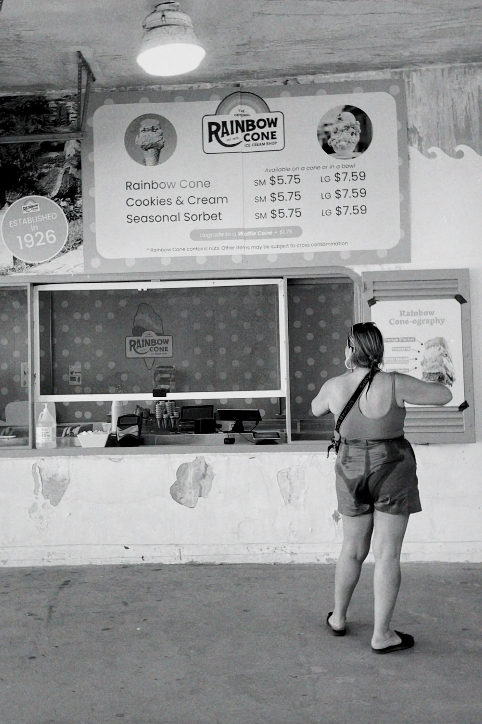 A woman photographer taking a photo of a Rainbow Cone ice cream stand at a beach, with a large sign displaying prices and flavors.