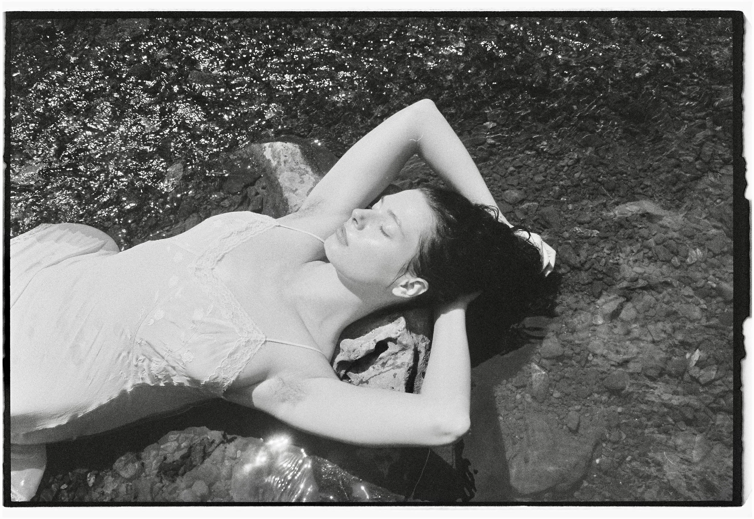 Black and white photograph of a woman lying on her back on a rocky surface with water nearby, her eyes closed and arms bent behind her head.