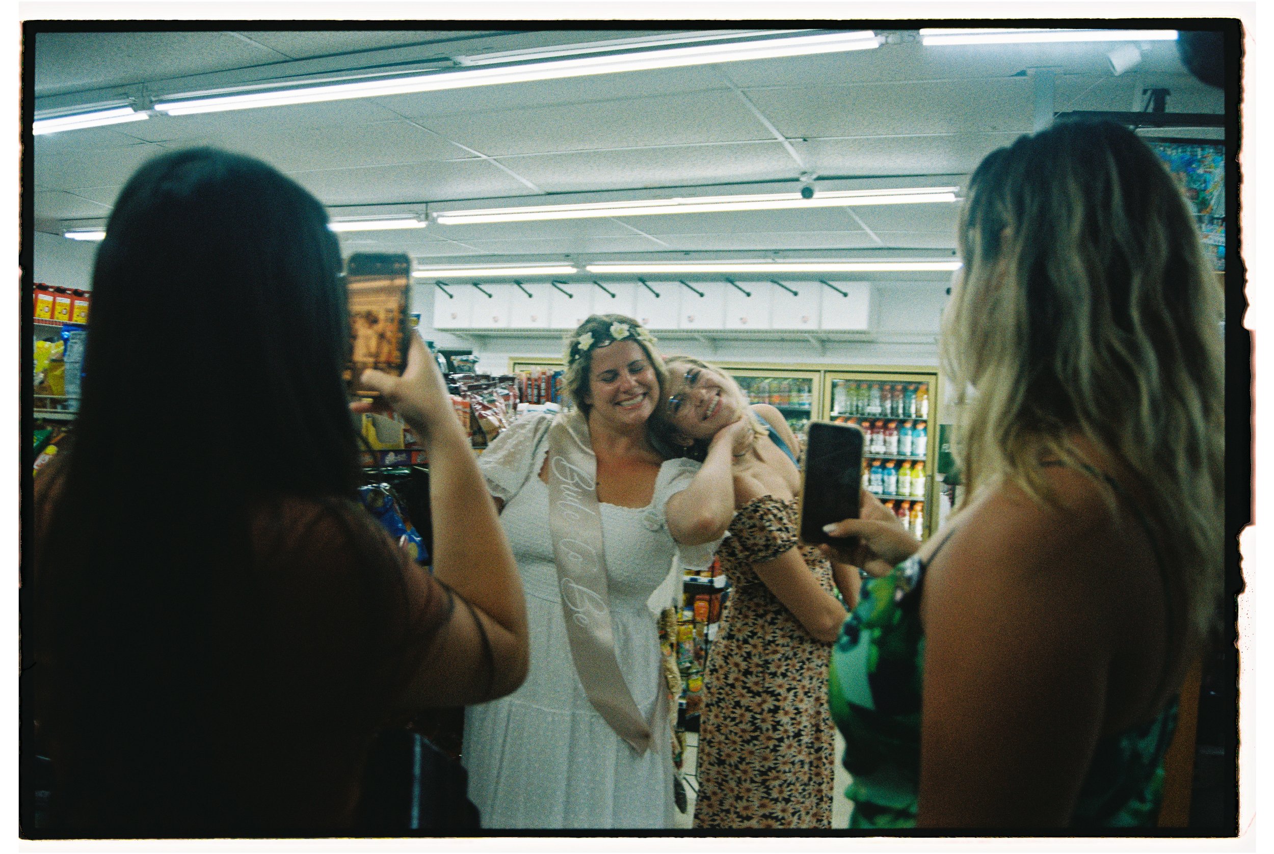 Four women in a grocery store, with one woman in a white dress and floral headband, wearing a sash that says 'Bride to Be,' taking and posing for photos with her friends.