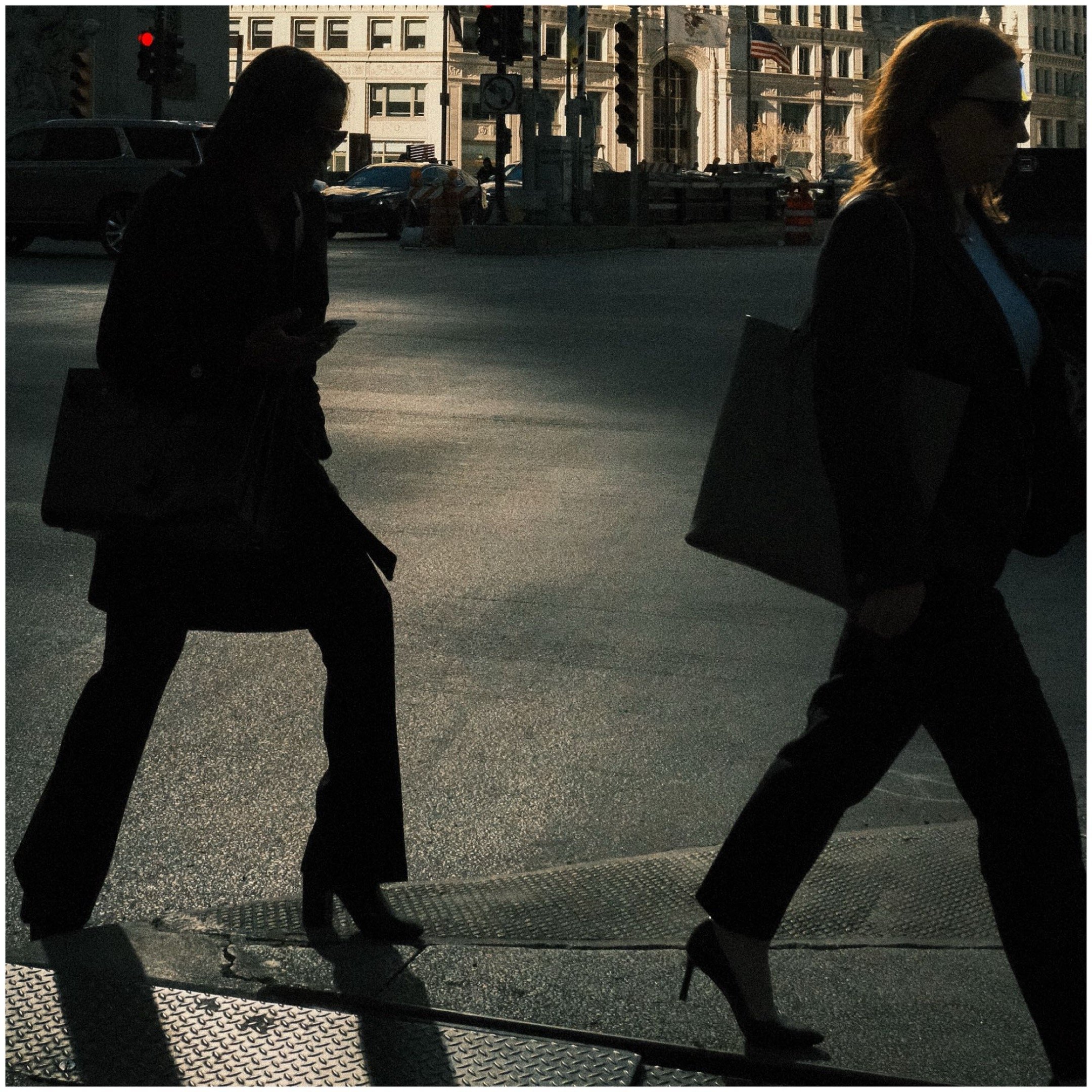 Two women walking across a city crosswalk, carrying handbags and wearing heels, with tall buildings and traffic lights in the background.