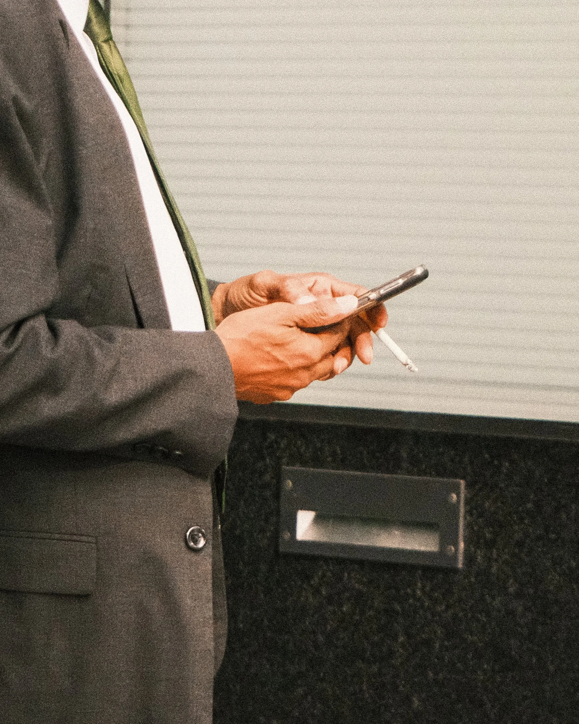 Close-up of a person in a suit holding a smartphone with a lit cigarette hanging from their fingers.
