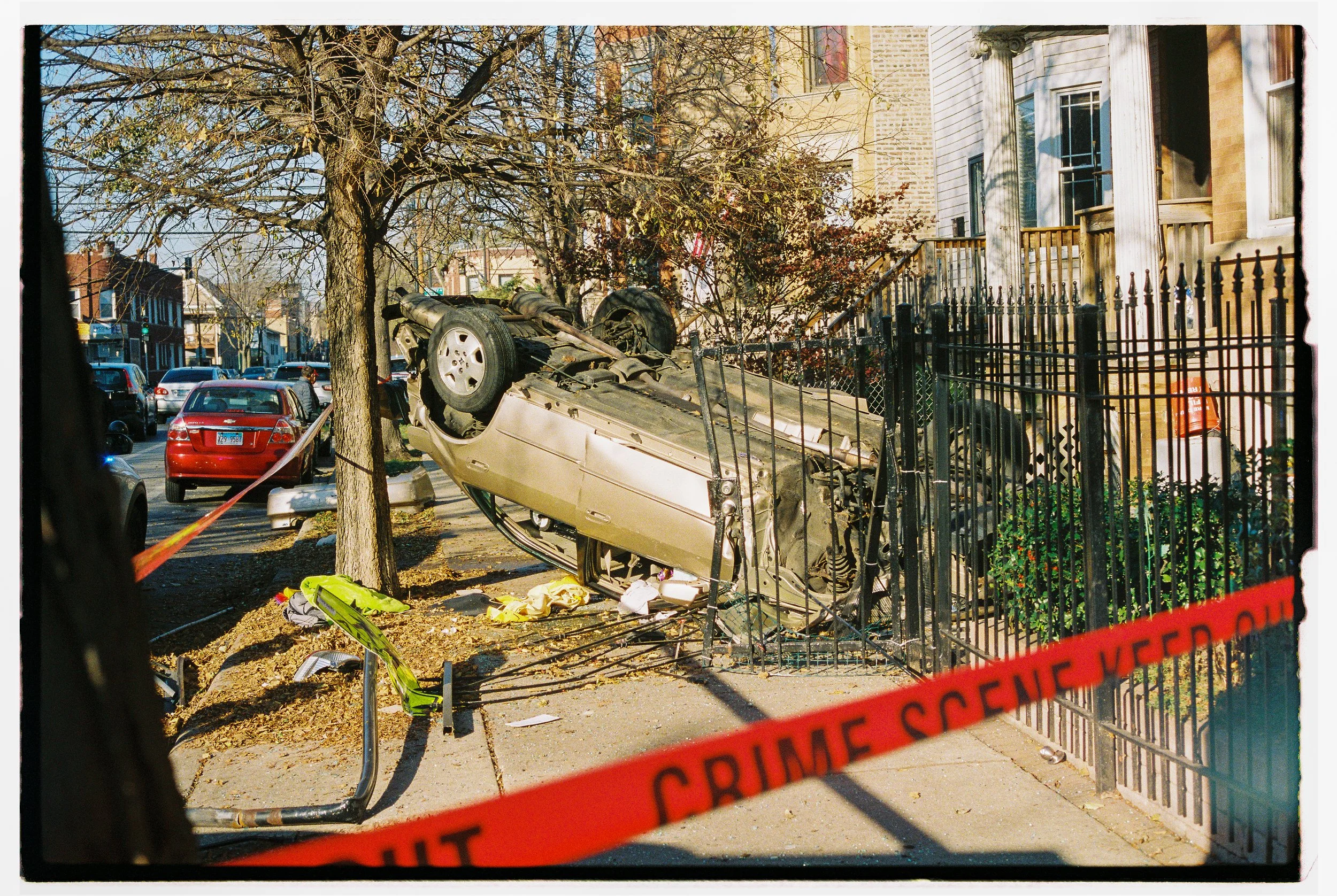 A car has overturned and crashed onto a sidewalk, hitting a metal fence and a tree. The vehicle is on its roof with debris scattered around. Police crime scene tape is visible across the scene.
