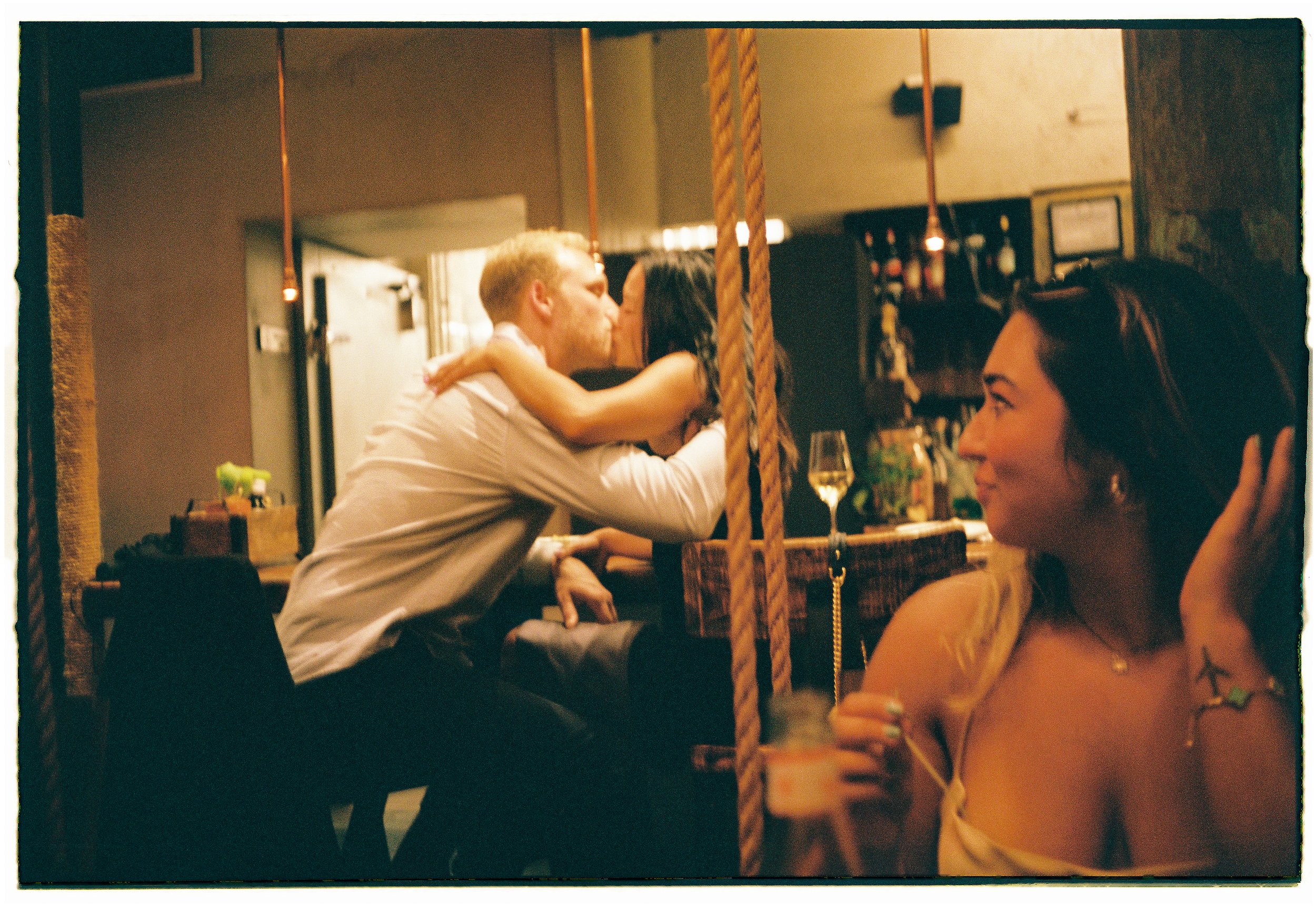 A couple kissing across a table at a bar or restaurant, with a woman sitting nearby in the foreground.