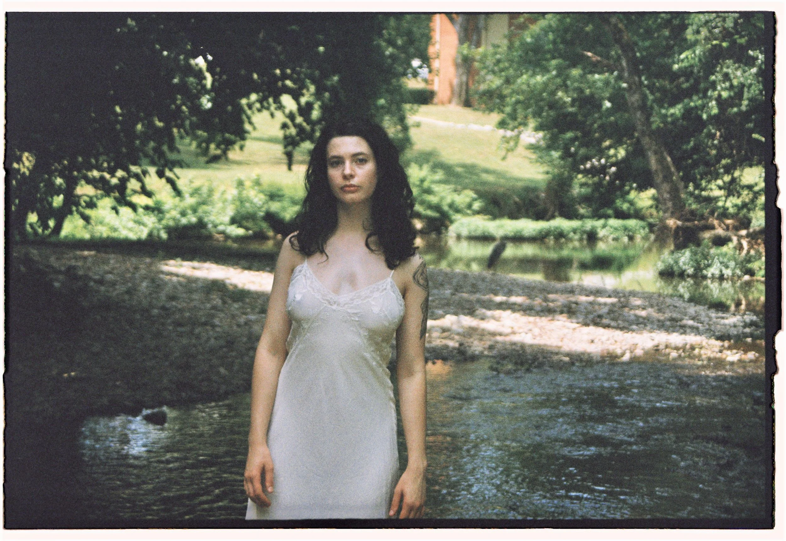 A woman with dark curly hair wearing a white dress standing near a creek in a park with green trees and grass in the background.