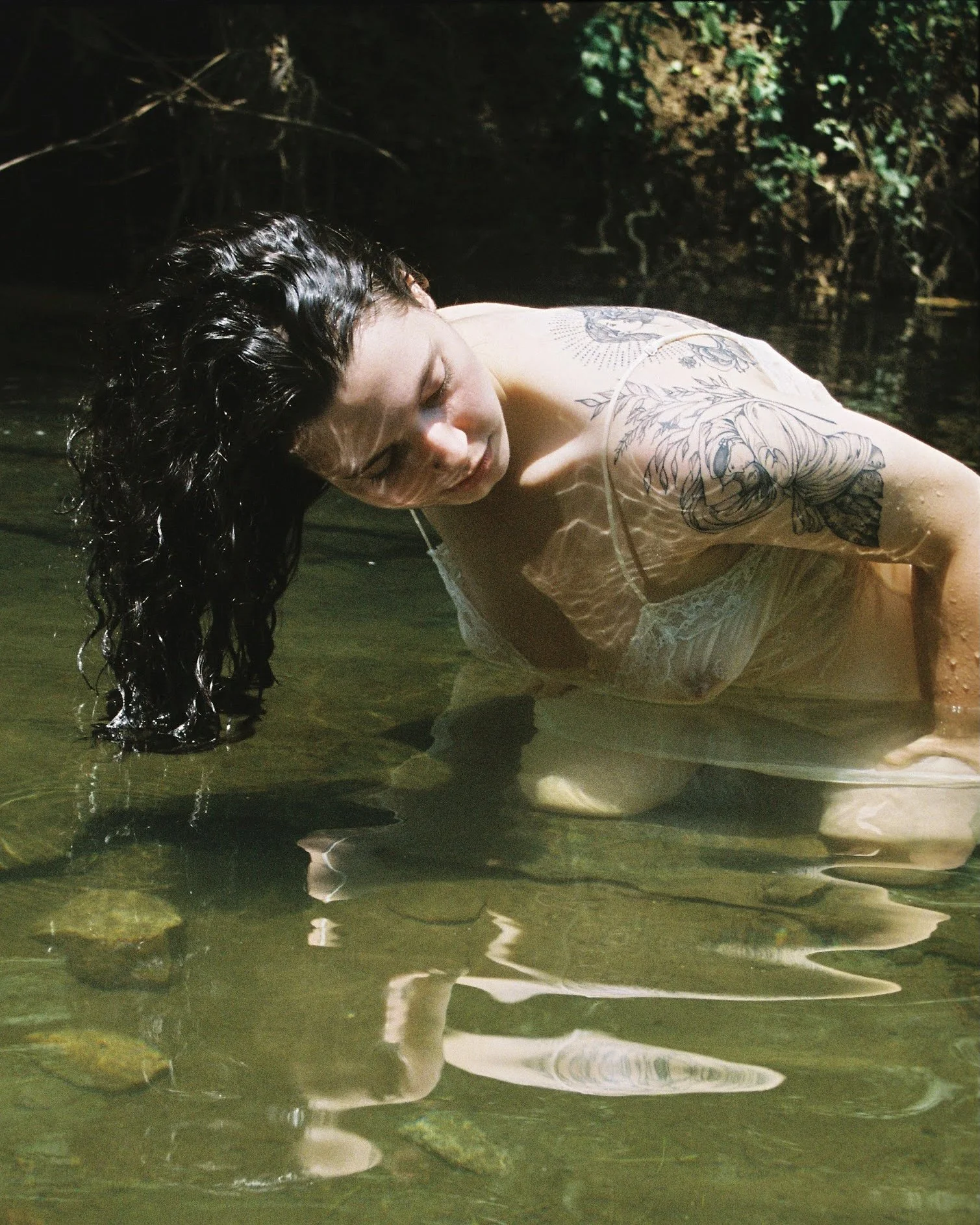 A woman with dark, curly hair and a tattoo on her arm, wearing a white lace dress, kneeling in a shallow creek with her head tilted down, water soaking her hair and dress, surrounded by green foliage.