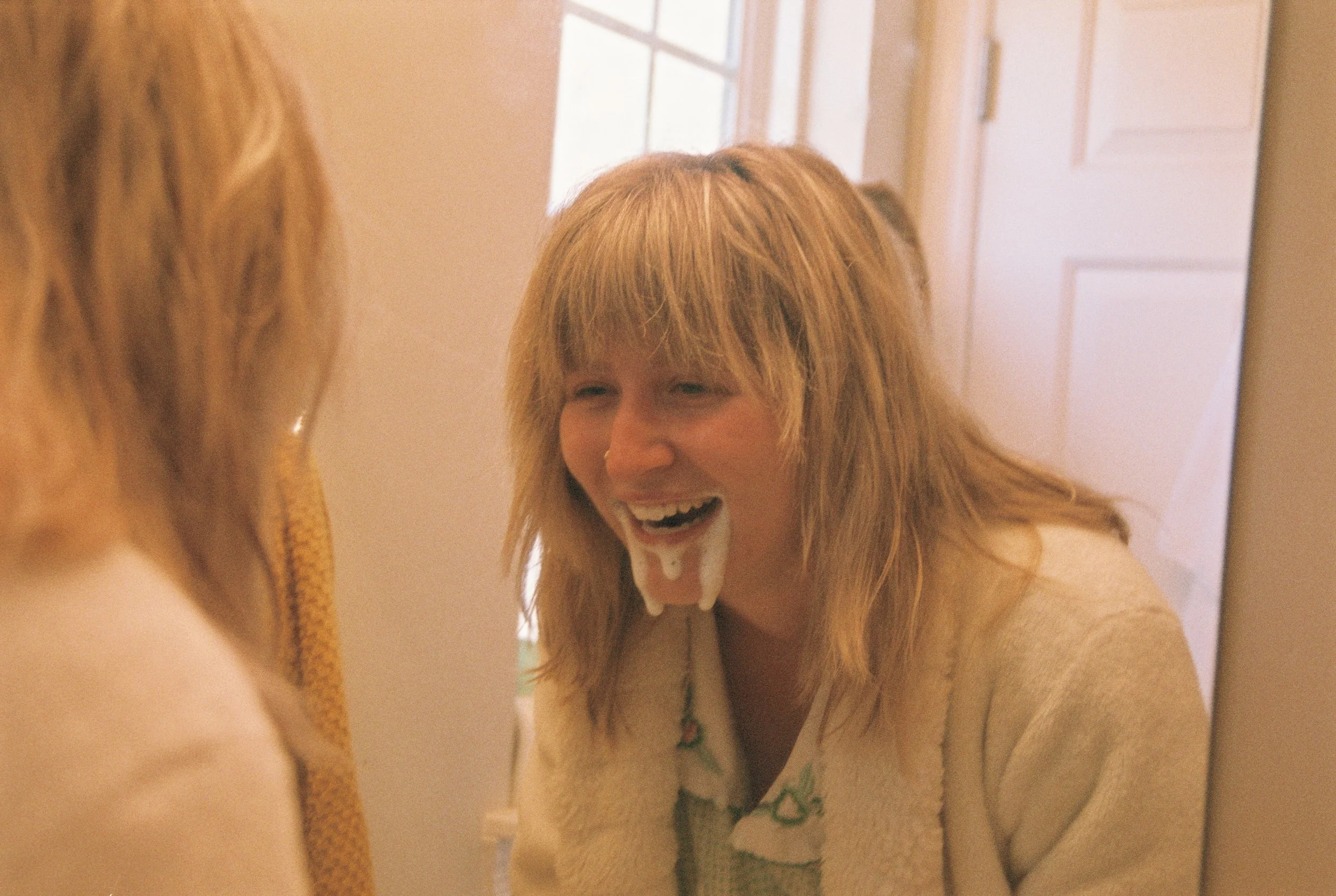 A woman with light brown hair looking into a mirror, laughing with foam on her chin.