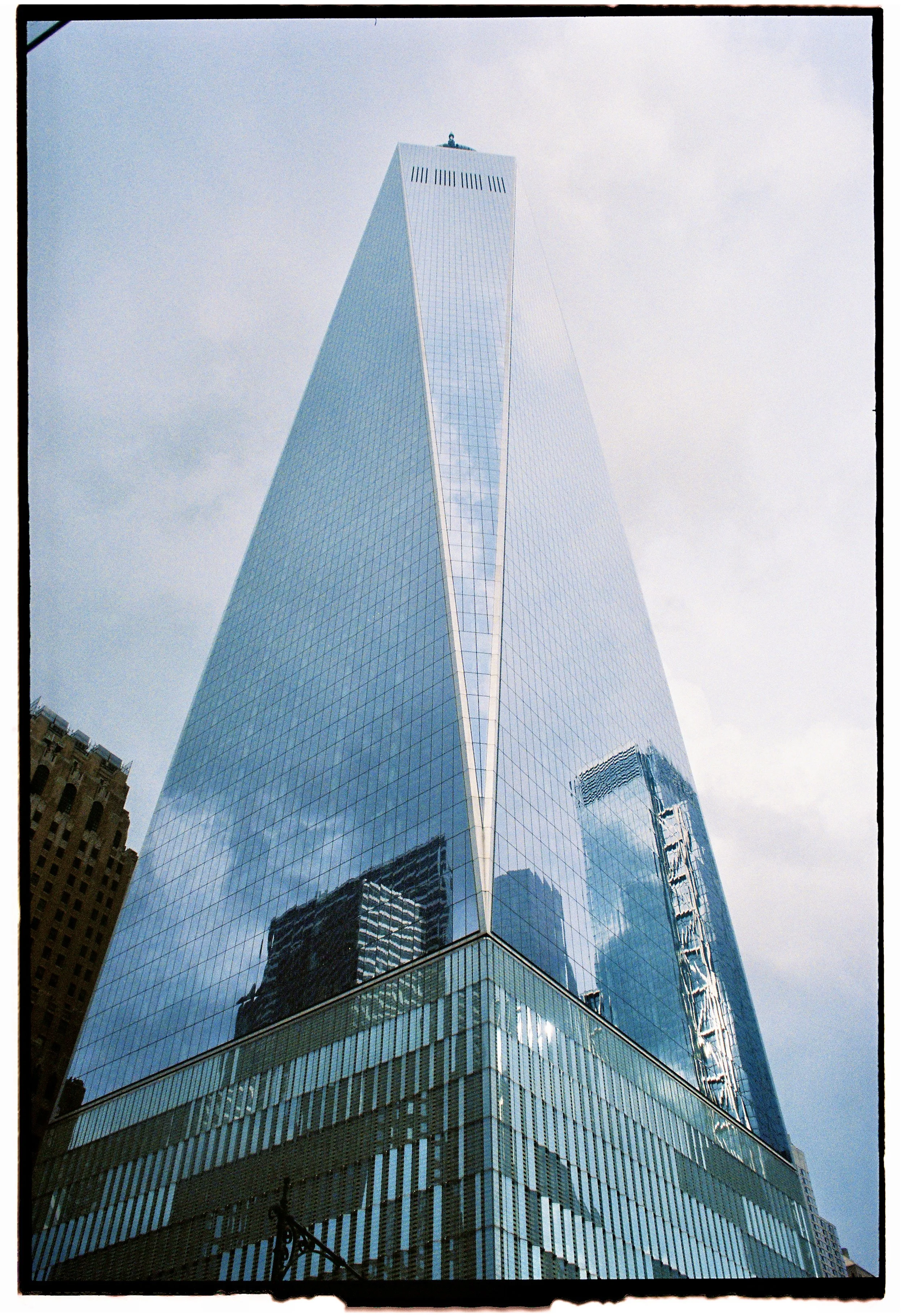 A tall glass skyscraper, the World Trade Center, with reflective windows, viewed from below, with clouds in the sky.