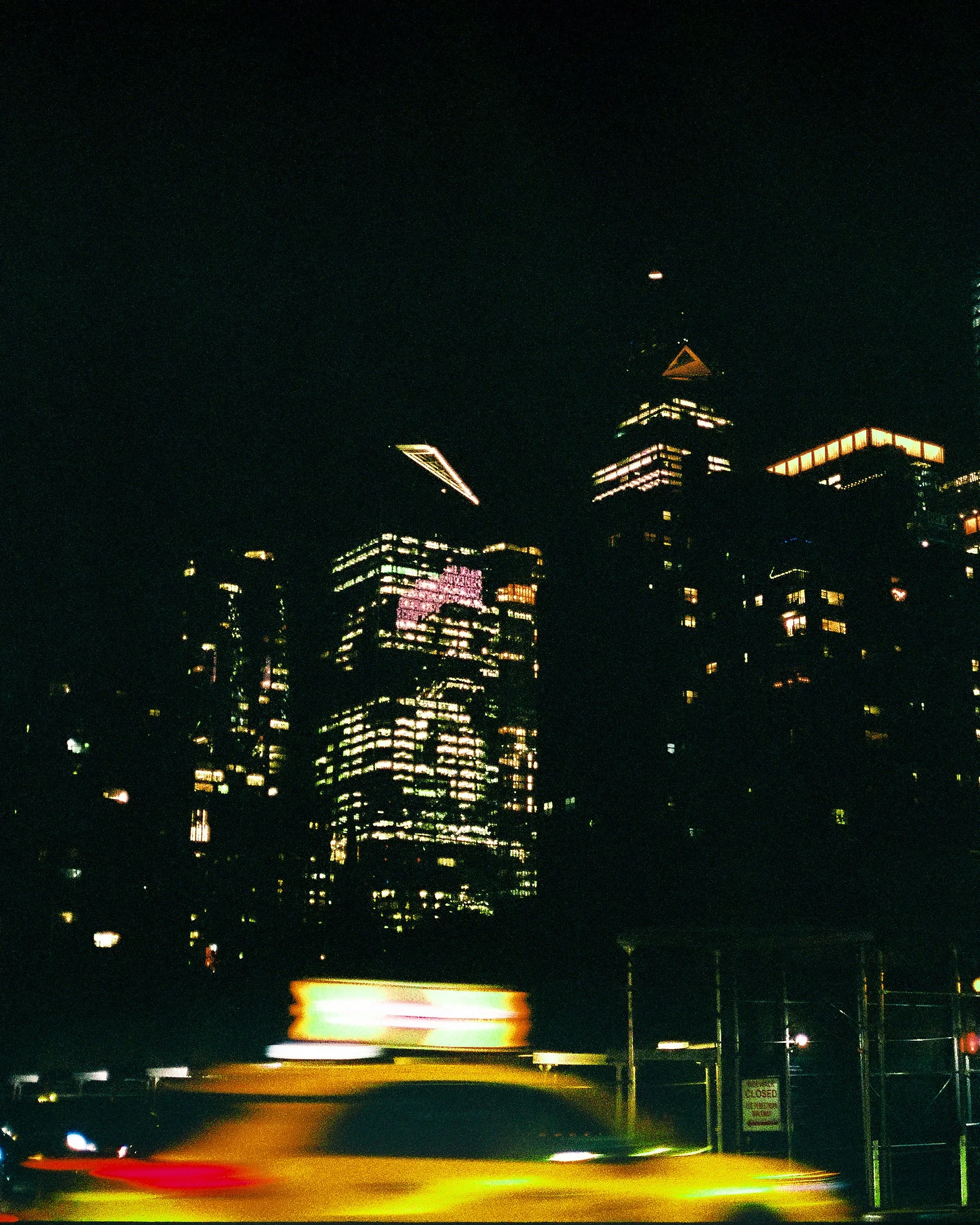 Nighttime cityscape with illuminated skyscrapers and a moving yellow taxi with a blurred sign in the foreground.