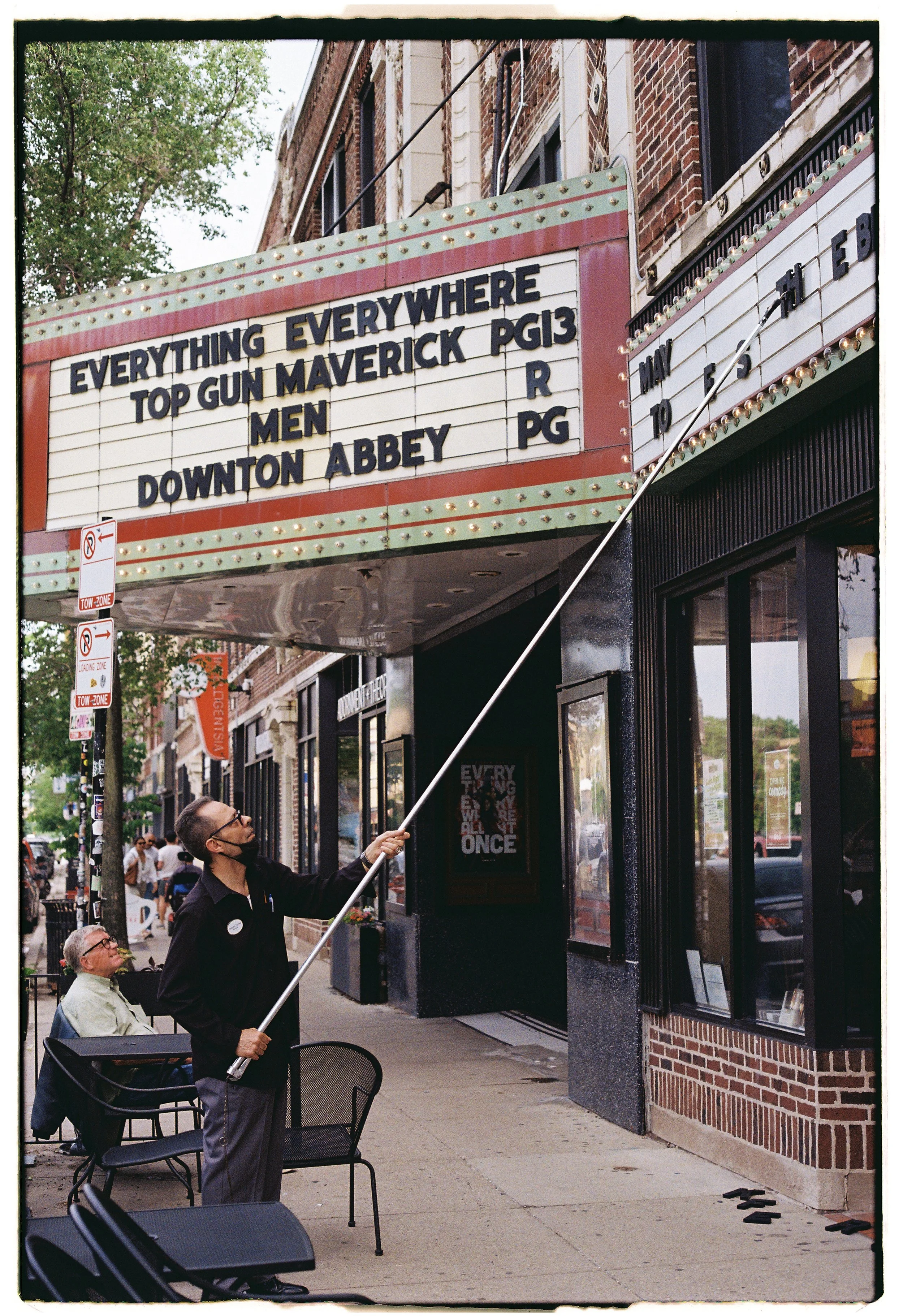 A man holding a pole on which a sign board is hanging, standing on a sidewalk outside a movie theater building. The sign board displays upcoming movie titles and ratings, including 'Top Gun Maverick PG13' and 'Downton Abbey PG. Behind him, another ma
