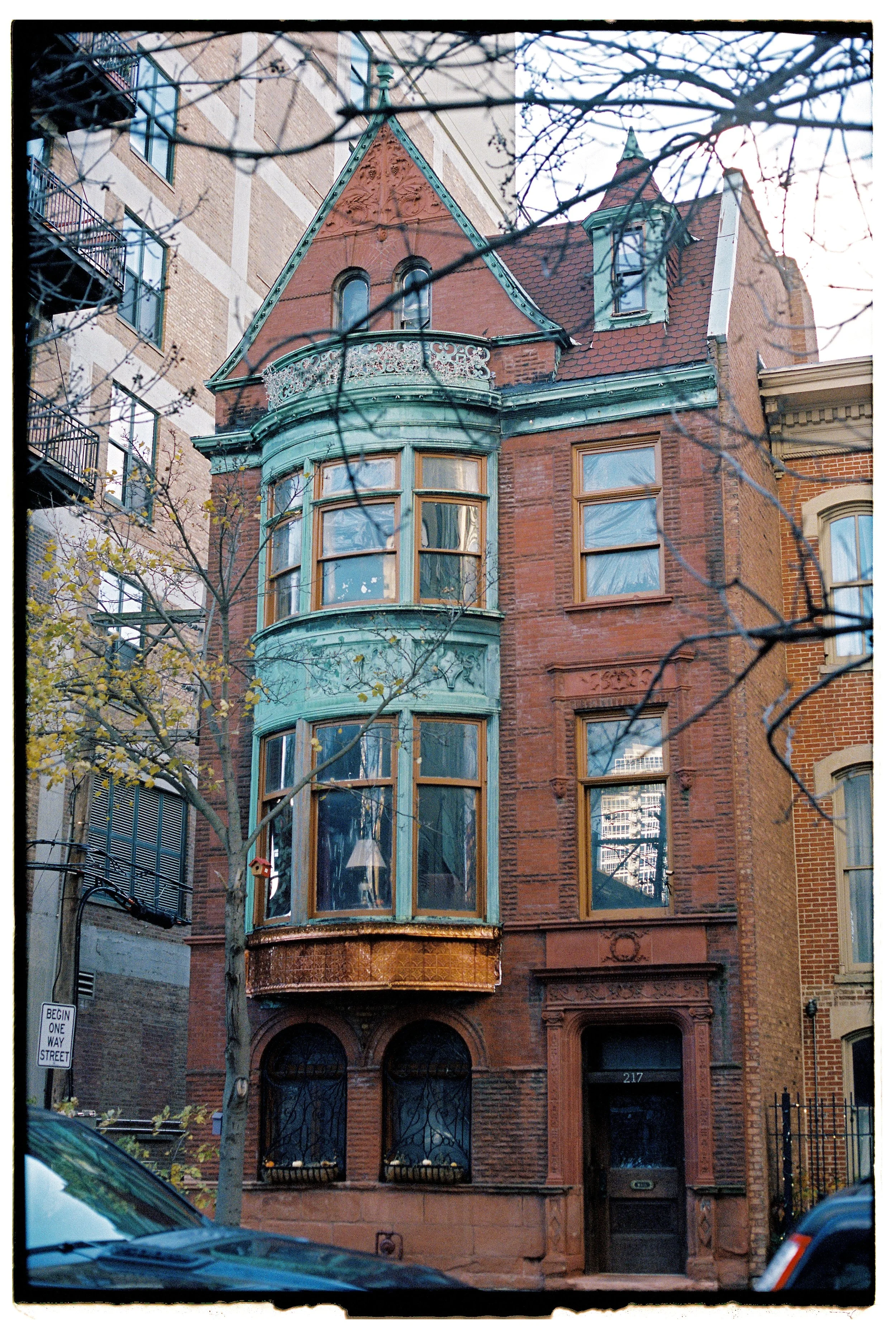 Historic red brick row house with a decorative green turret and bay window, situated between taller buildings, with a tree and parked cars in front.
