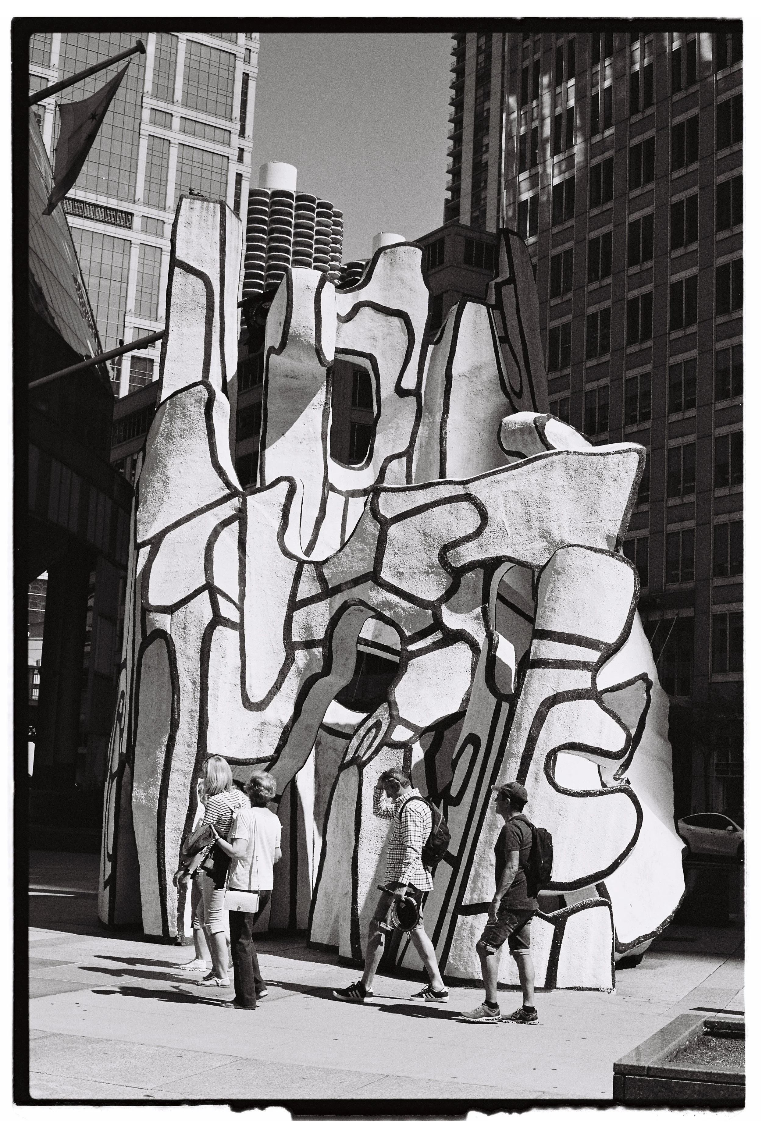 People walking past a large abstract sculpture with geometric shapes and black lines, set against tall buildings in an urban downtown area.