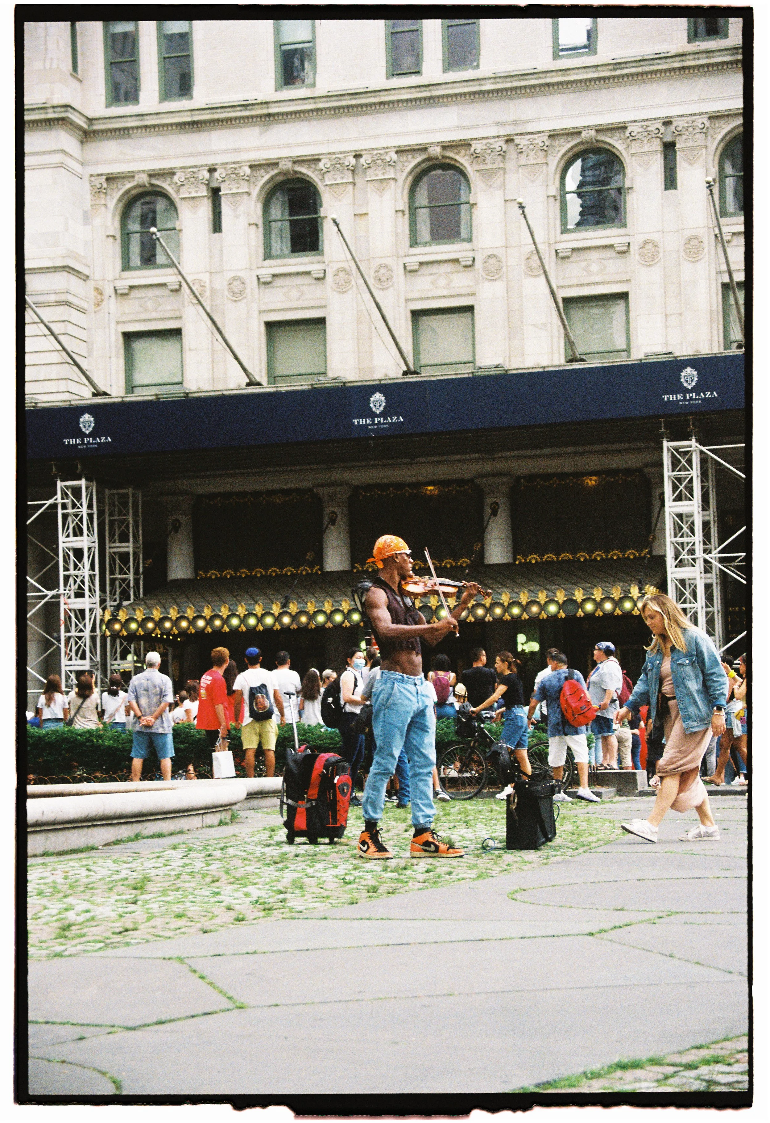 A street performer playing a violin in front of a crowd in New York City, with a large building and an awning labeled 'The Plaza' in the background.