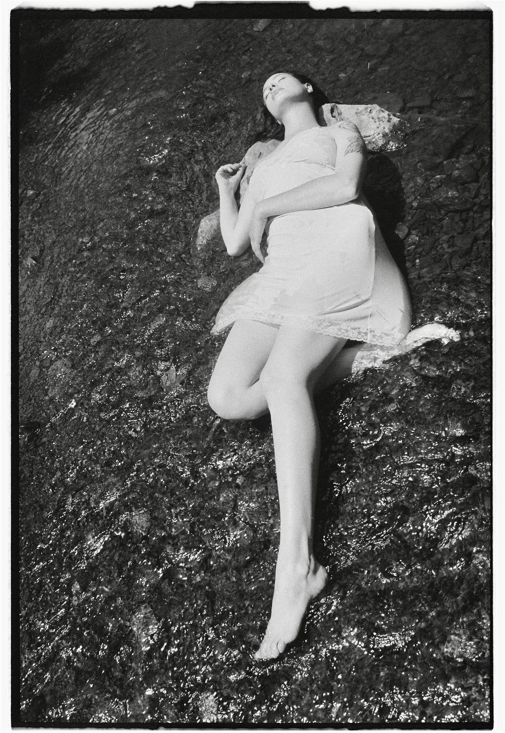 A woman lying on a large rock near water, wearing a sleeveless dress with her eyes closed.