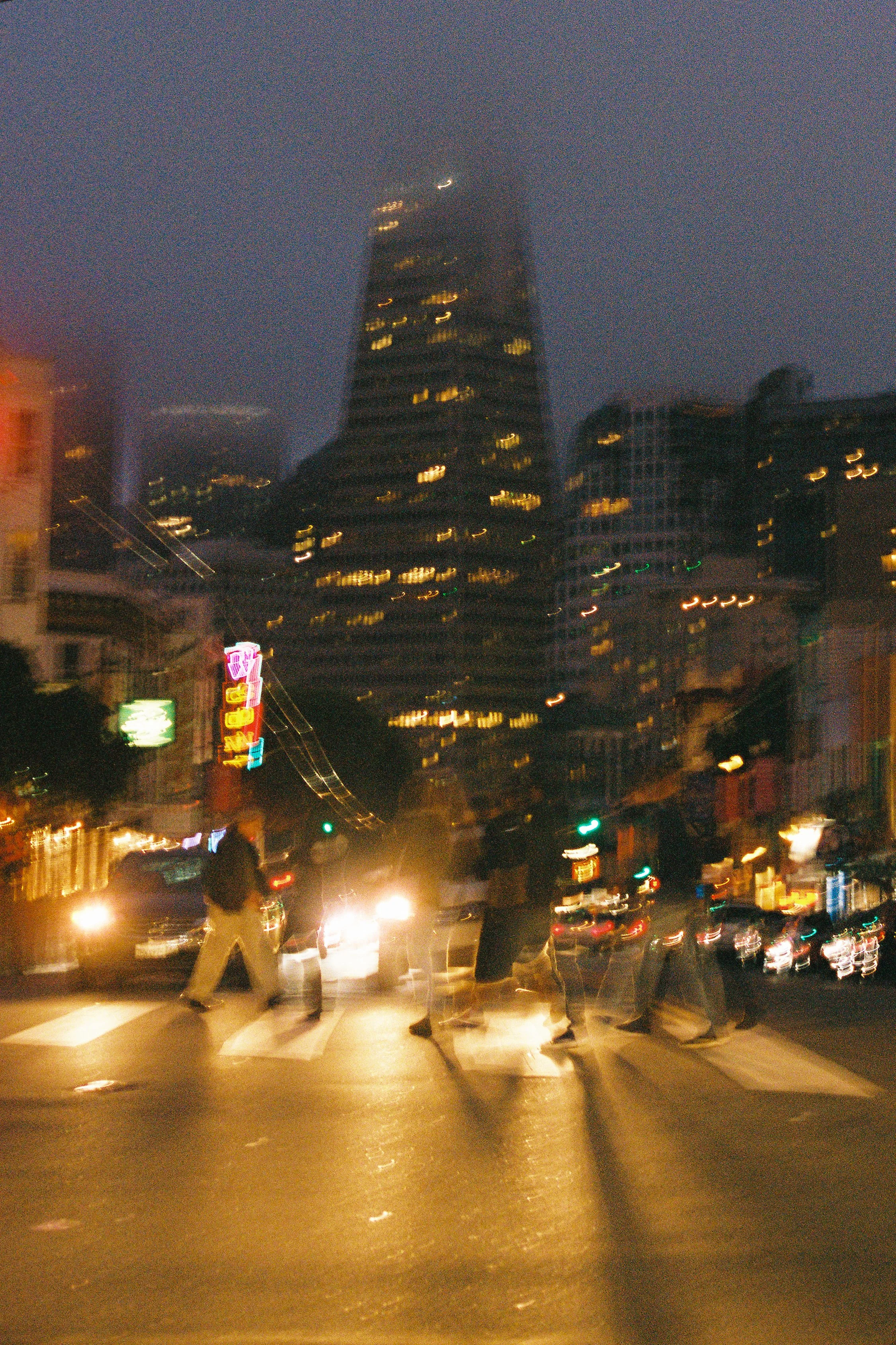 Nighttime city street with blurry pedestrians crossing a crosswalk and a tall building with illuminated windows in the background.