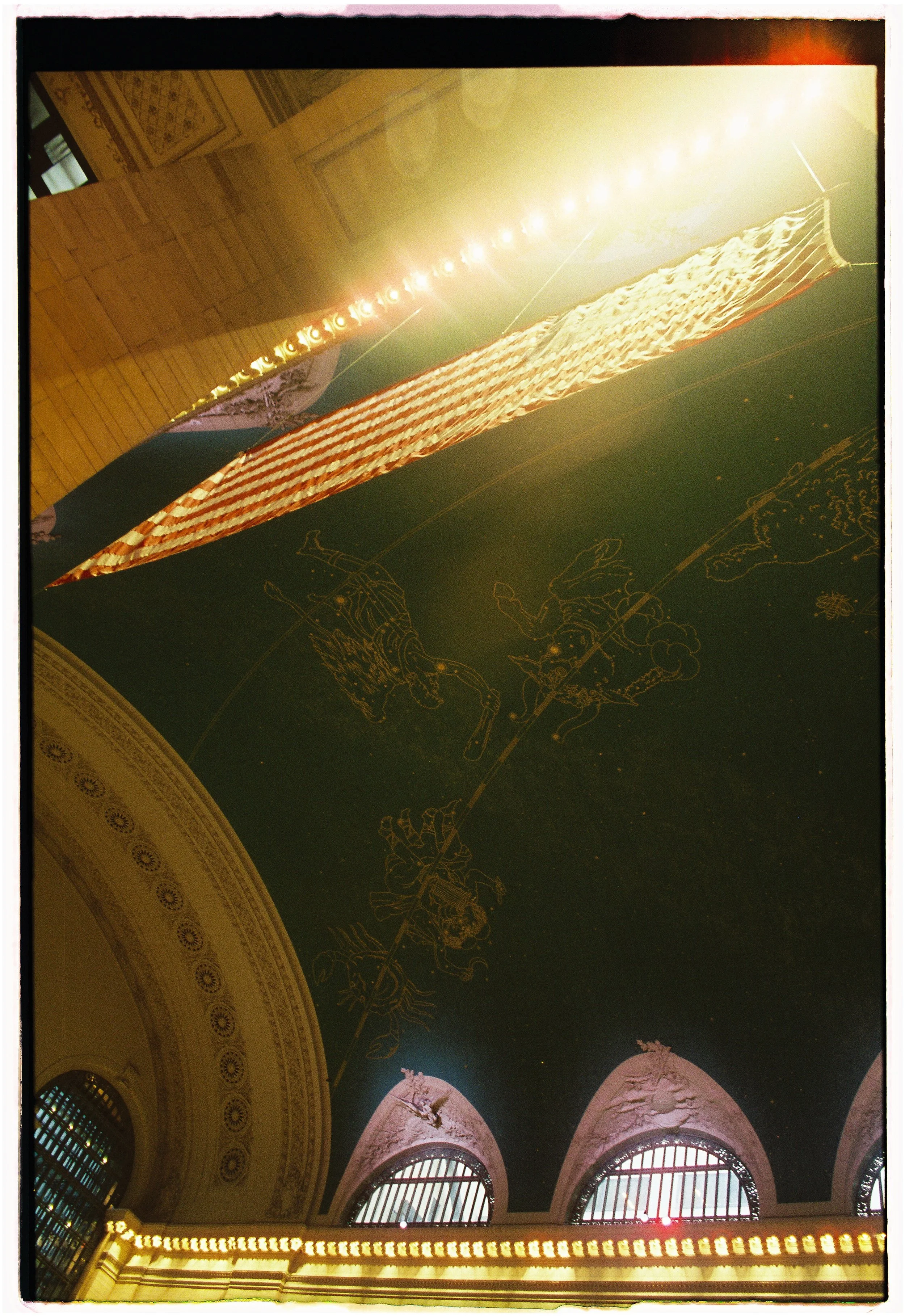 The image shows an ornate ceiling decorated with a constellation of zodiac signs, with some stars and a sunlit glowing effect. There are four arched windows with barred grilles and decorative molding below the ceiling.