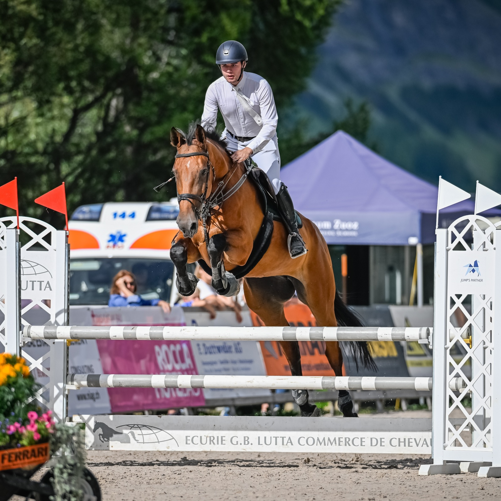 Reiter springt mit seinem Pferd über einen Hindernis im Voltigier- oder Springreiten. Es trägt einen Reithelm und Reitkleidung. Im Hintergrund sind Bäume, Zuschauer und Zelt zu sehen.