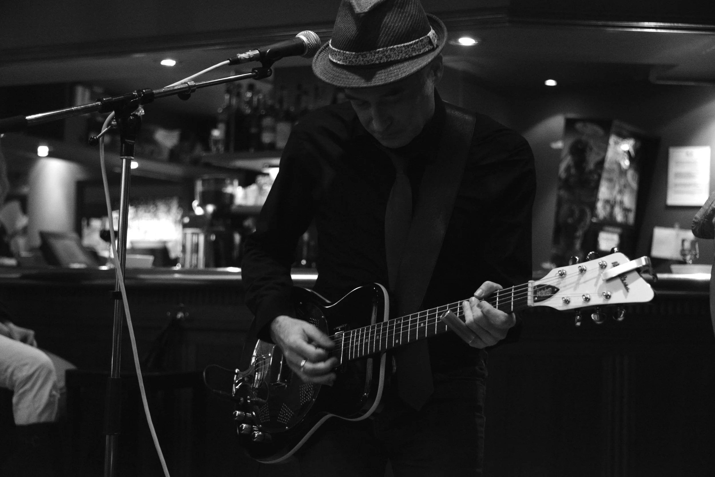 A musician playing an electric guitar in a dimly lit bar or club, wearing a hat and dark clothing, with a microphone stand in front of him.