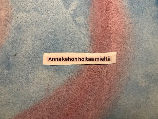 Typed note in Finnish saying 'Anna kehon hoitaa mieltä' on a textured outdoor wall.