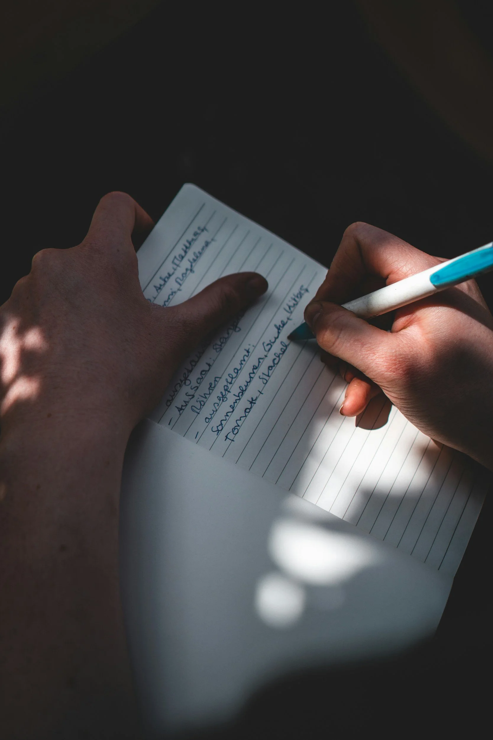 Close-up of hands writing in a lined notebook with a blue pen, with shadows cast across the page.