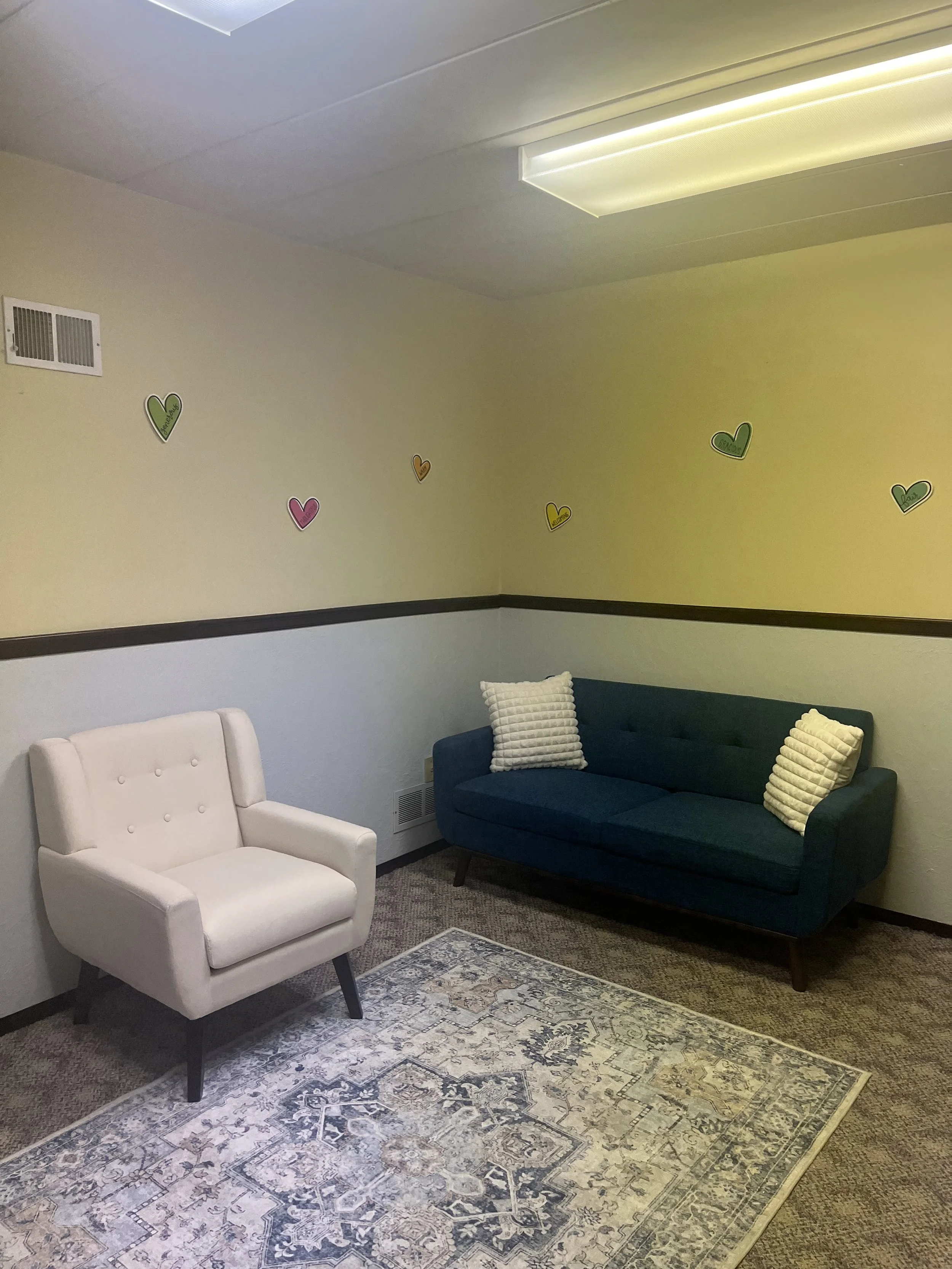 A sitting room with a white armchair, a dark blue sofa with two off-white pillows, a patterned rug, and decorated with colorful heart-shaped wall stickers on the yellow painted wall.