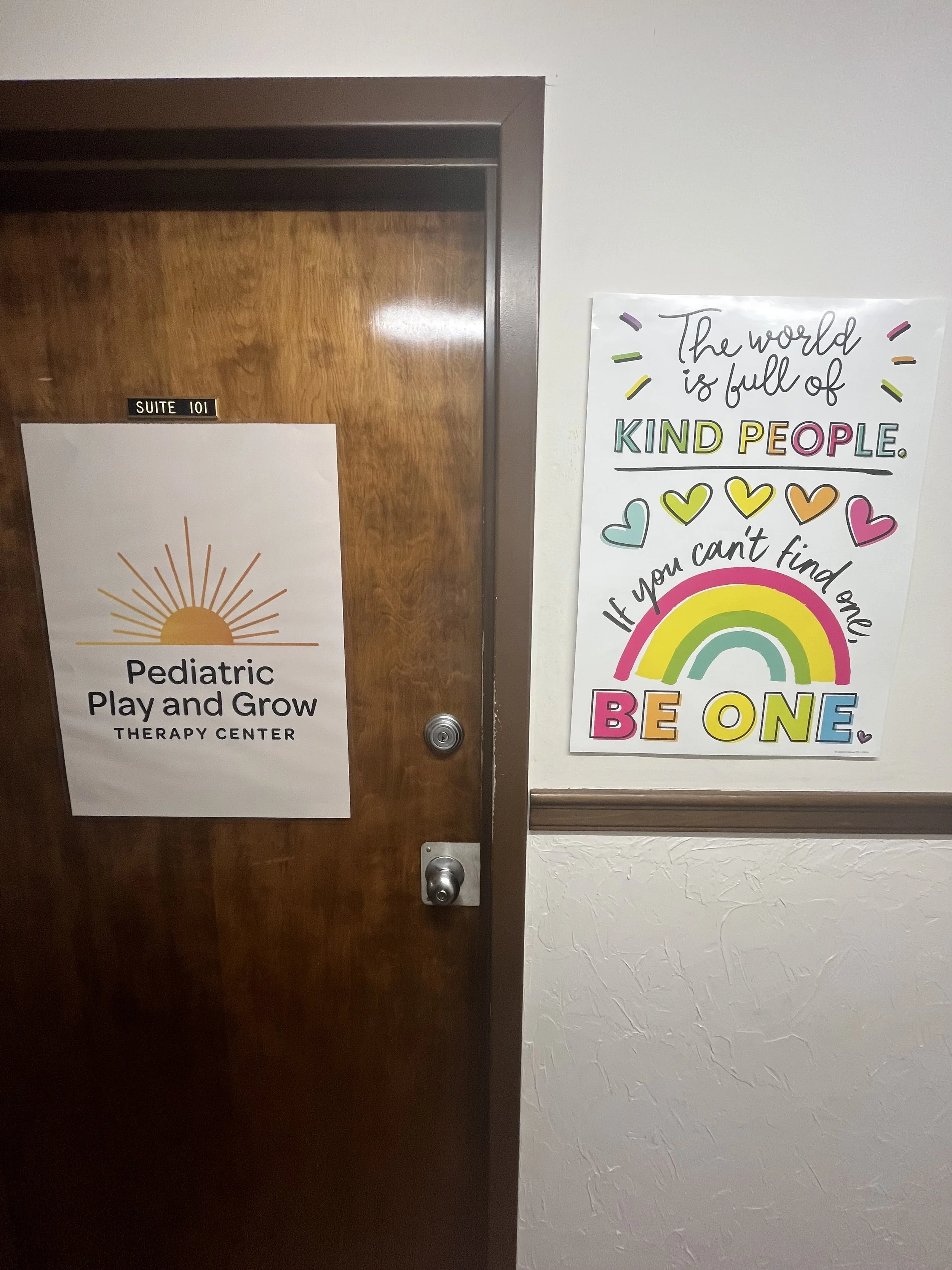 A wooden door labeled Suite 101 with a sign that reads 'Pediatric Play and Grow Therapy Center' and a colorful poster on the wall beside it with inspirational words and a rainbow.