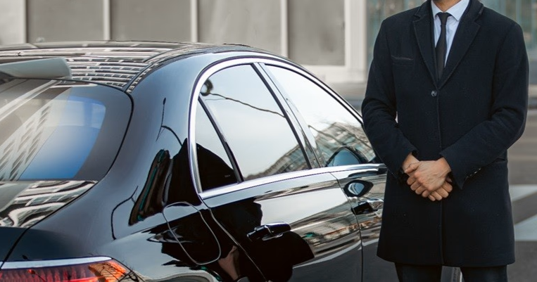 A private chauffeur from Black Car Agency in a suit standing next to a Black Car Mercedes S-Class.