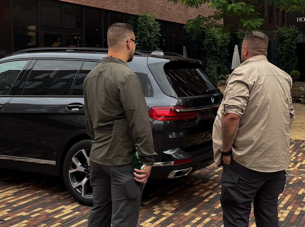 Two men standing outdoors near a black BMW SUV having a conversation, with trees and a building in the background.