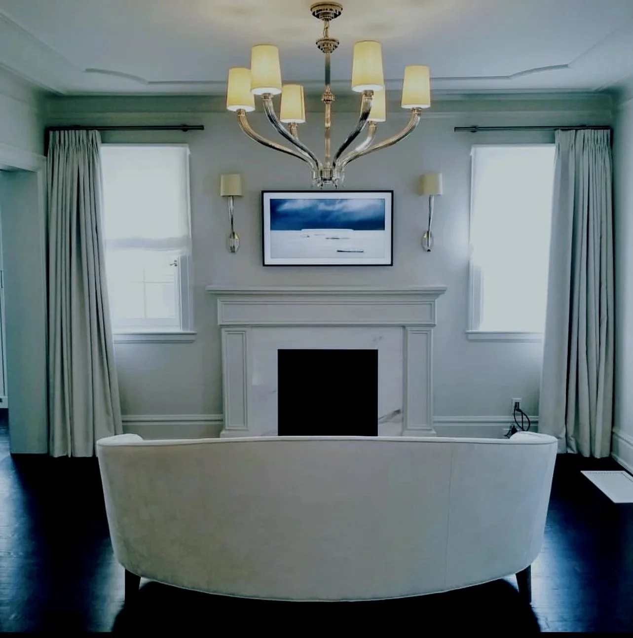Living room with white walls, curtains, and a fireplace with a framed picture above it. There is a chandelier hanging from the ceiling and a cream-colored sofa facing the fireplace. Two windows with white curtains flank the fireplace.
