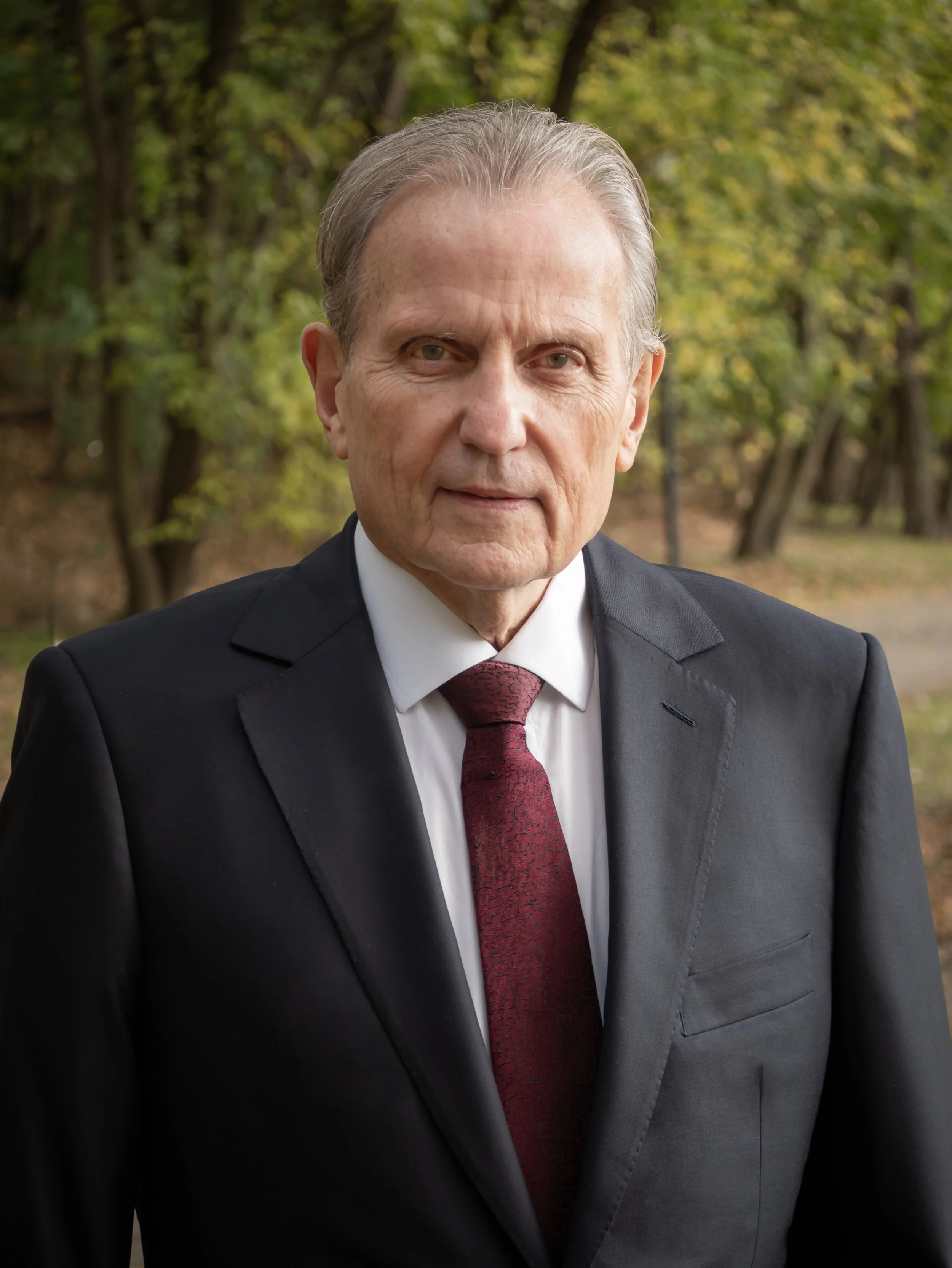 A mature man in a black suit, white shirt, and red tie standing outdoors with trees in the background.