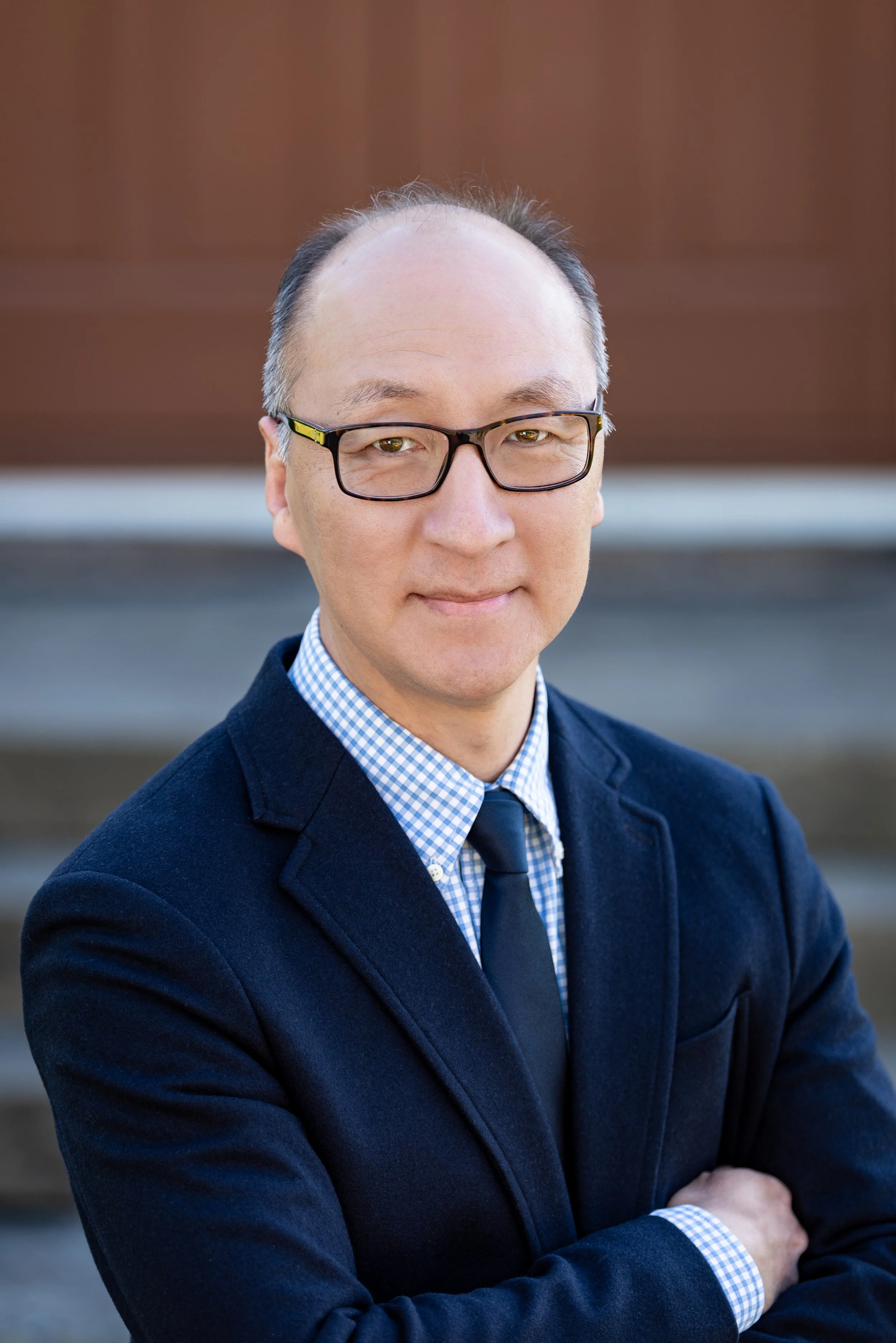 A man wearing glasses, a navy blue blazer, a light blue checkered shirt, and a navy tie, standing outdoors with arms crossed, smiling gently, with a blurred background of brown and gray tones.