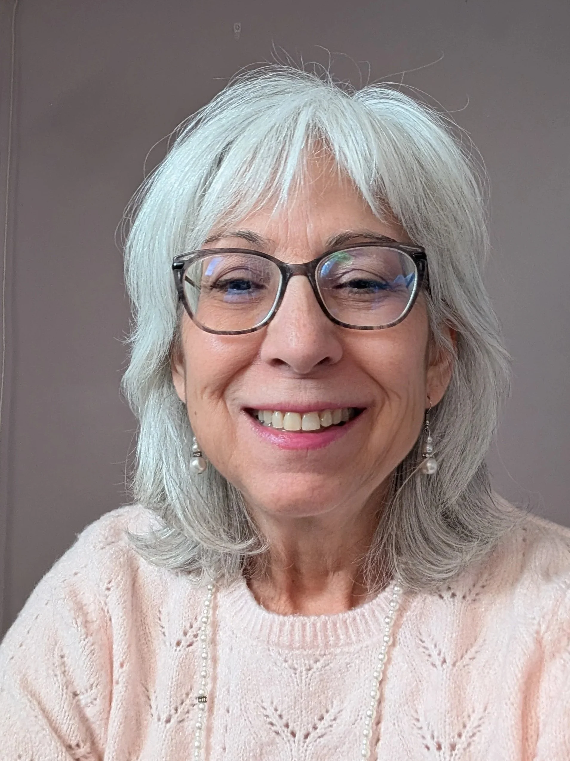 Une femme, avec de beaux cheveux blancs, vêtue d'un pull rose pâle, des boucles d'oreilles perlées et un collier de perles, souriant dans un décor intérieur simple.