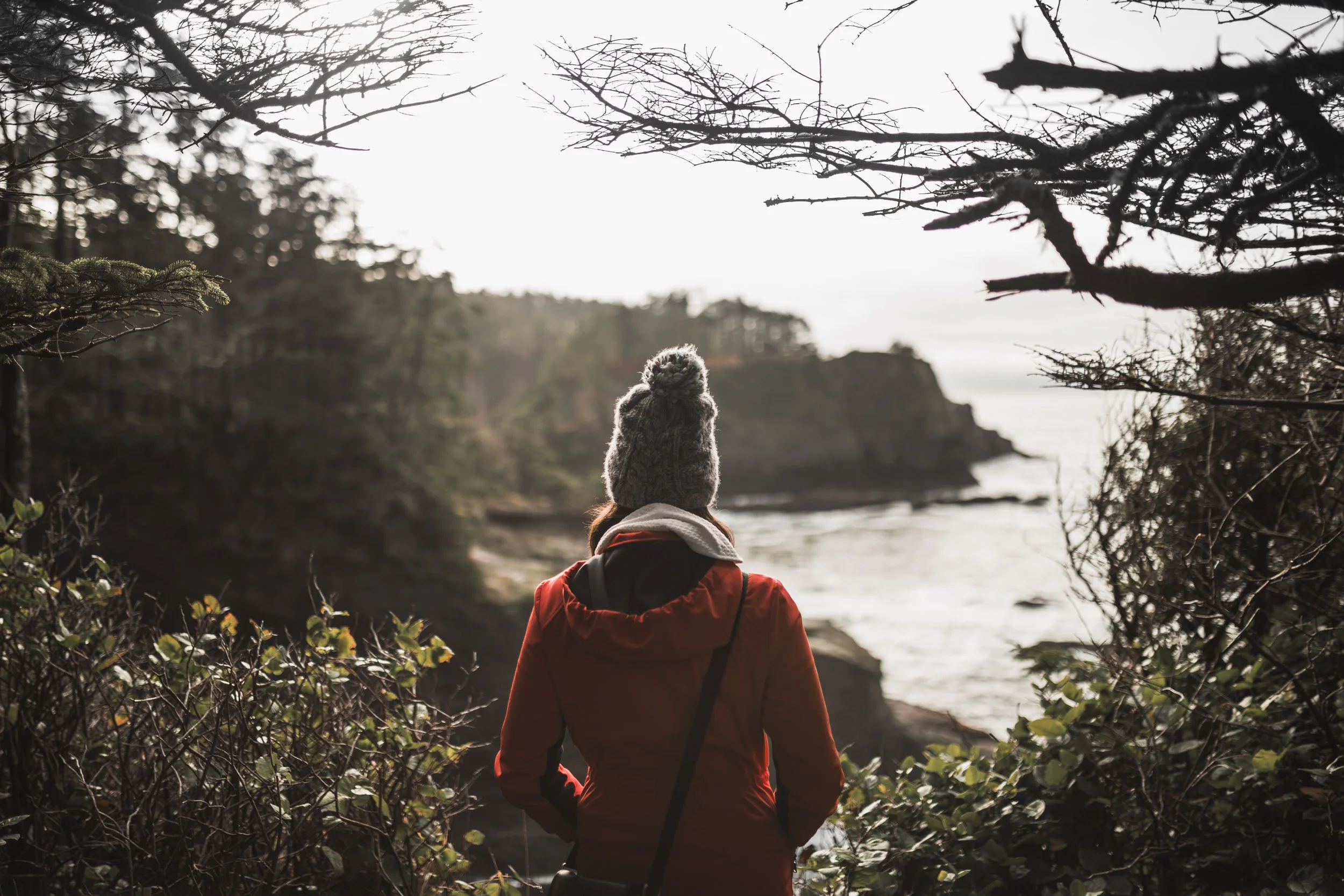 Personne vue de dos, portant un manteau rouge et un bonnet en laine grise, regardant la mer depuis une forêt sur une côte rocheuse.