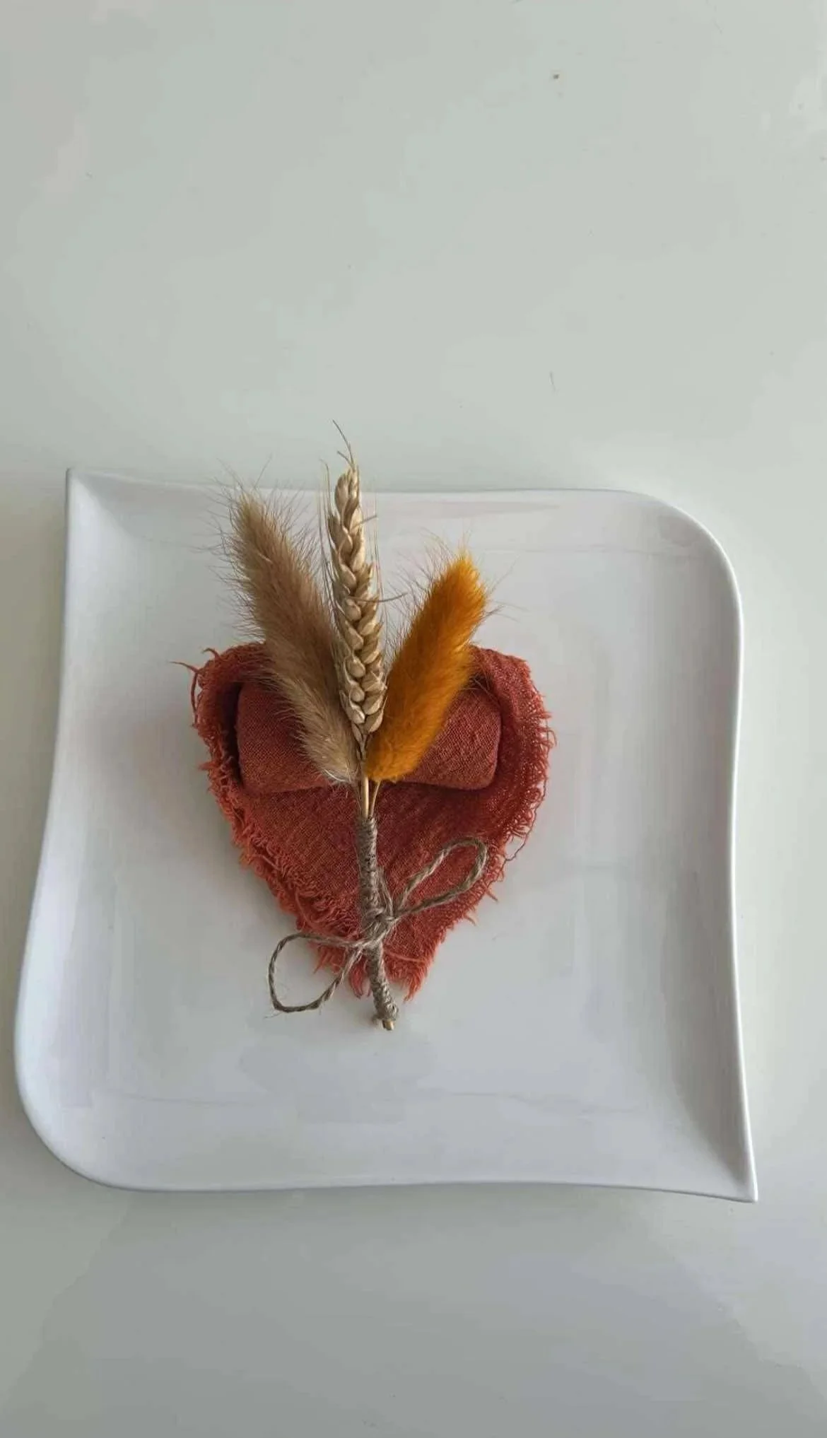 Une décoration composée d'une branche de blé, de plumes de différentes couleurs, attachée à un cœur en tissu rouge, placé sur une assiette blanche.