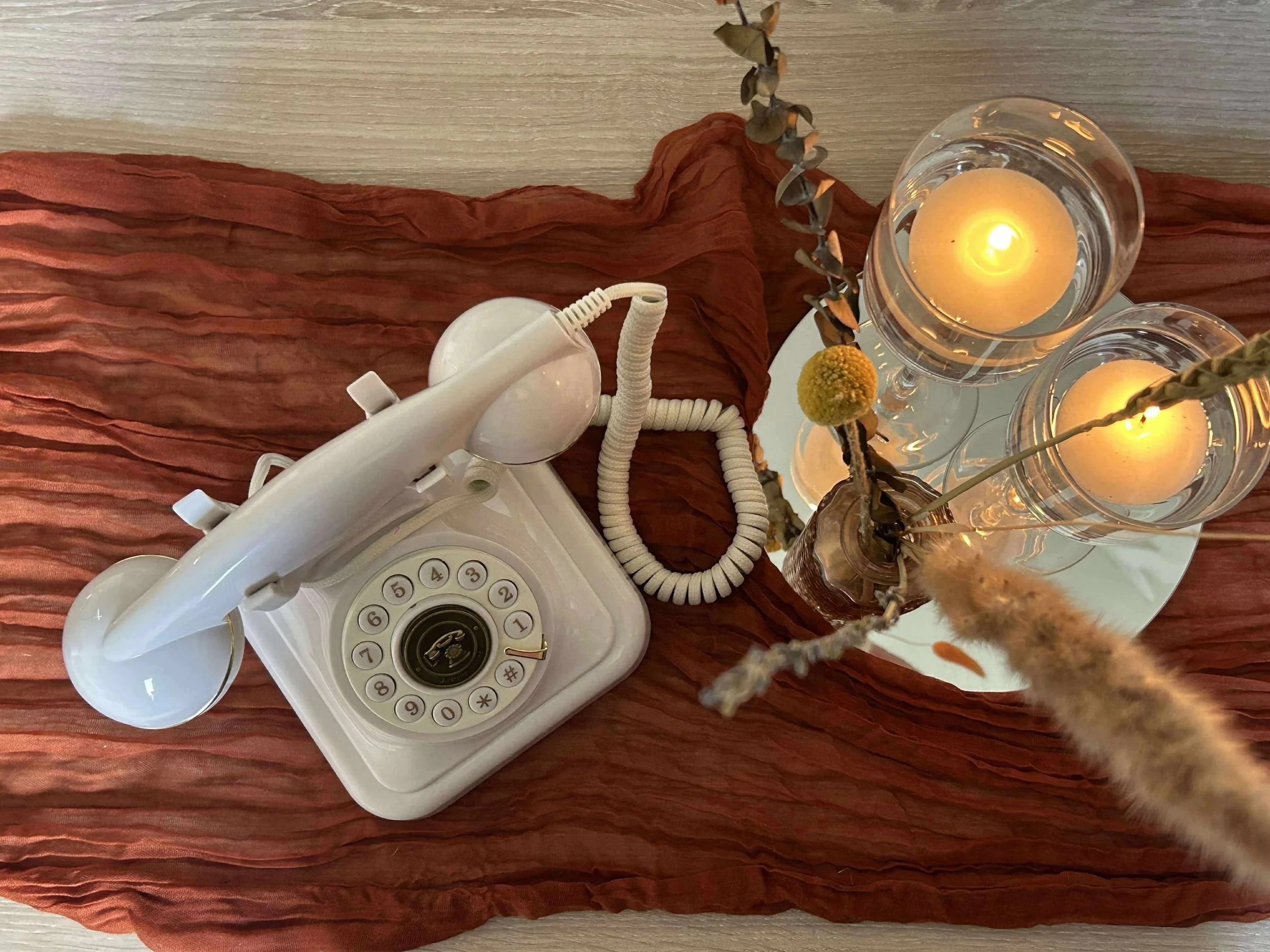Un téléphone vintage blanc sur un tapis de couleur cuivrée, avec une affiche de fleurs séchées dans un vase en verre et deux bougies allumées dans des verres à pied sur une surface en bois.