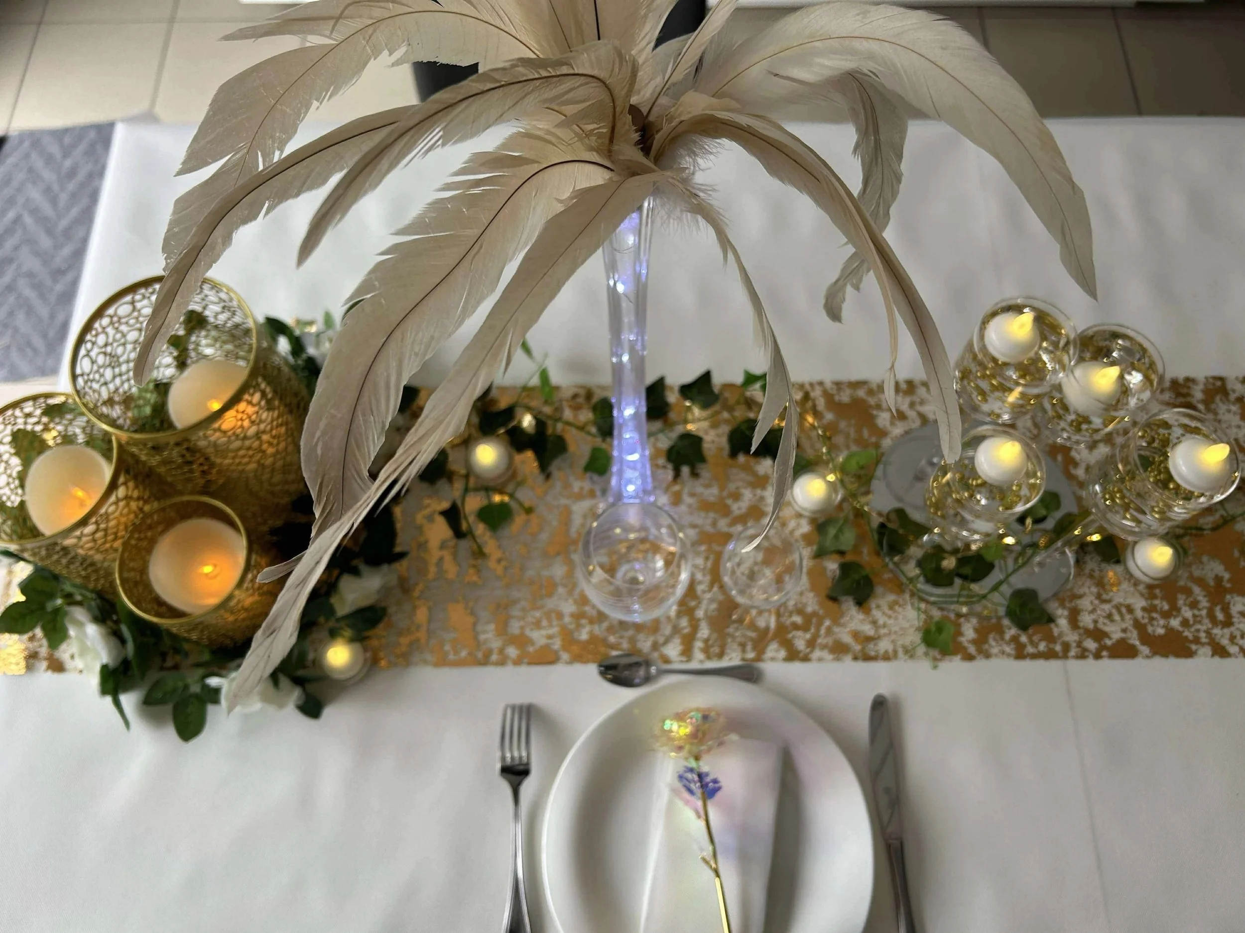 Table décorative avec des bougies, des plumes blanches, des bougeoirs dorés, des lanternes en verre avec des bougies, une guirlande de feuillage, et une assiette avec des couverts.
