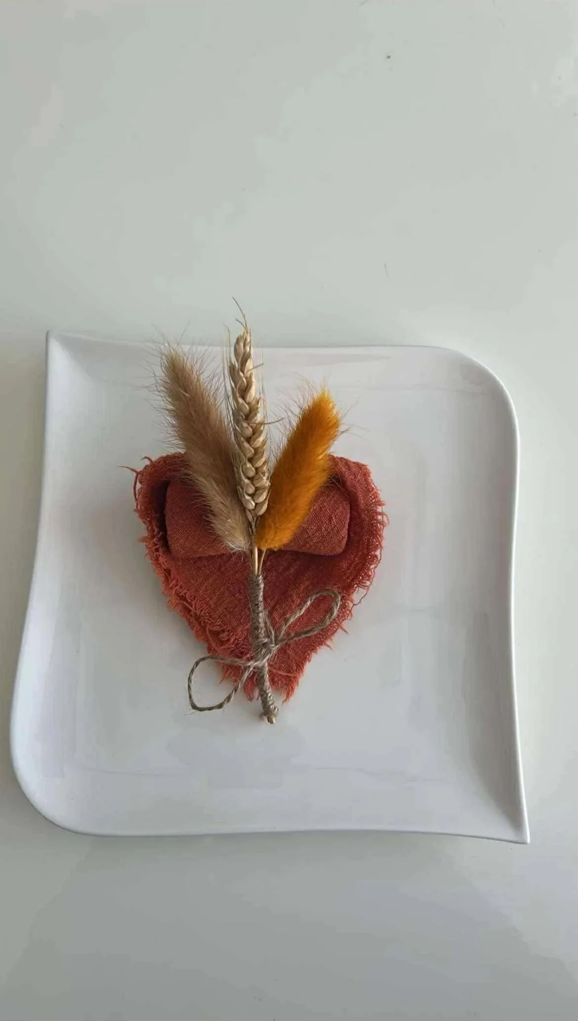 Un cœur en tissu rouge avec des plumes naturelles et orange attachées avec une ficelle, placé sur une assiette blanche