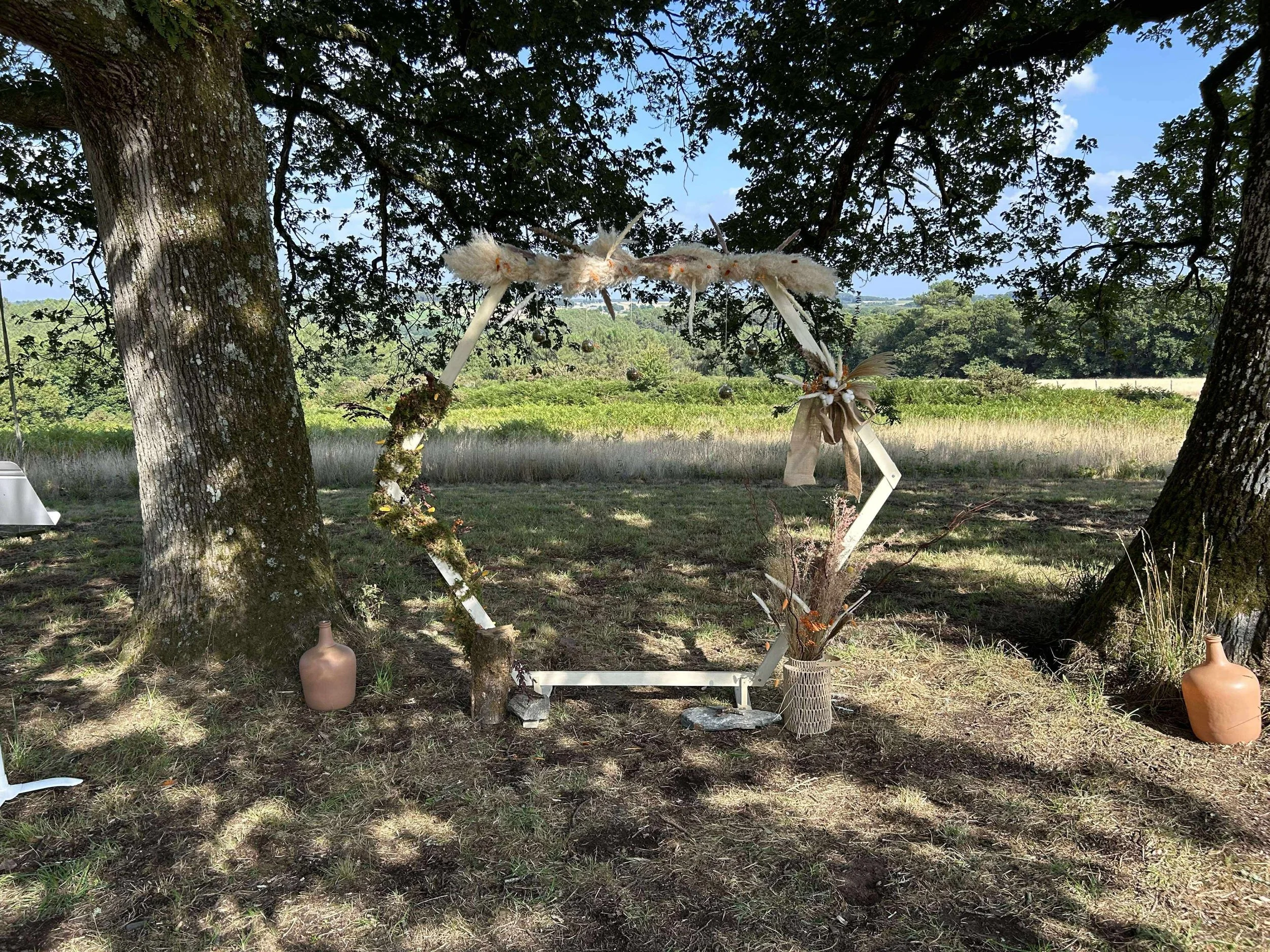 Un cadre décoratif en forme de cercle suspendu entre deux arbres, décoré de fleurs, de branches, de rubans et de textiles, avec des vases à ses pieds dans un champ herbeux sous un ciel bleu.