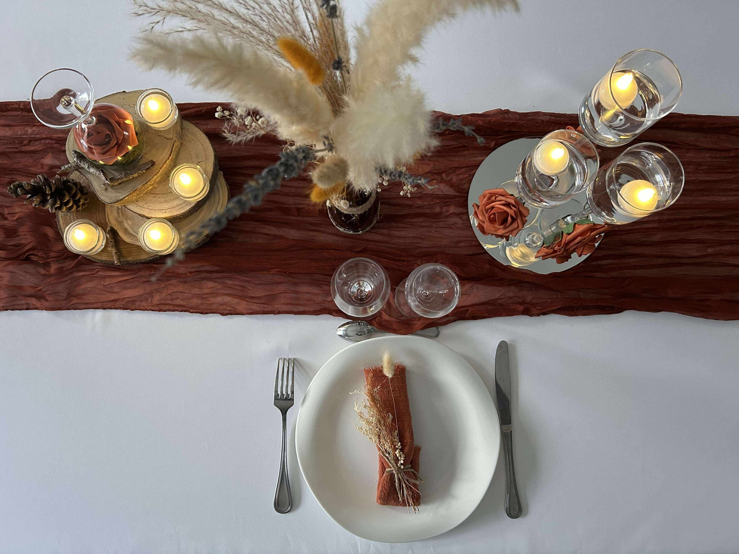 Une table élégante avec une nappe blanche, un centre de table avec des bougies, des fleurs en rouge et beige et des éléments décoratifs en bois et en laine. Présentoirs avec des bougeoirs en verre contenant des bougies allumées, et un endroit pour manger avec une assiette, un couteau, une fourchette et un tissus marron avec des fleurs et une corde.